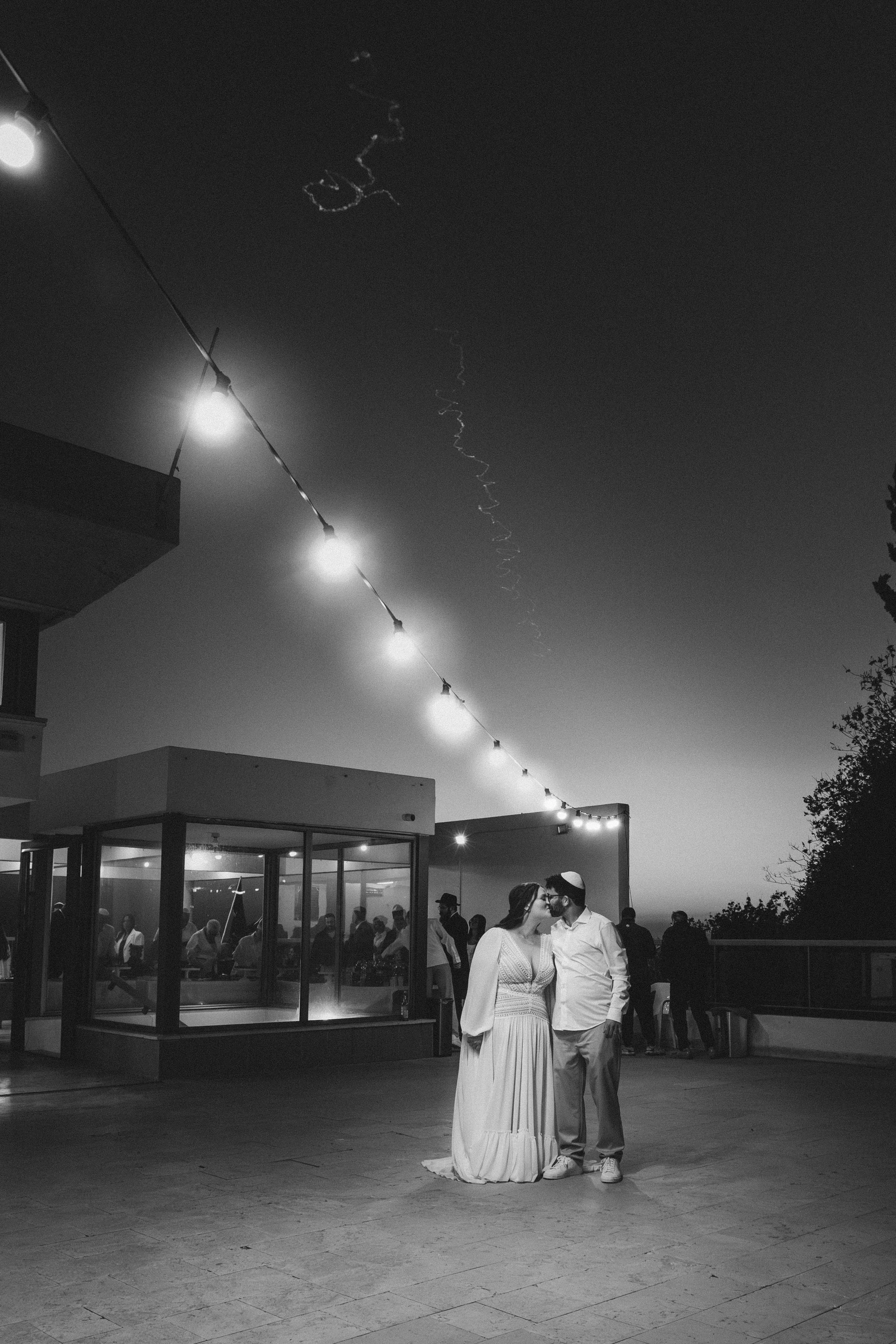 A bride and groom kissing at an outdoor wedding reception during the evening, with guests in the background and string lights hanging overhead.