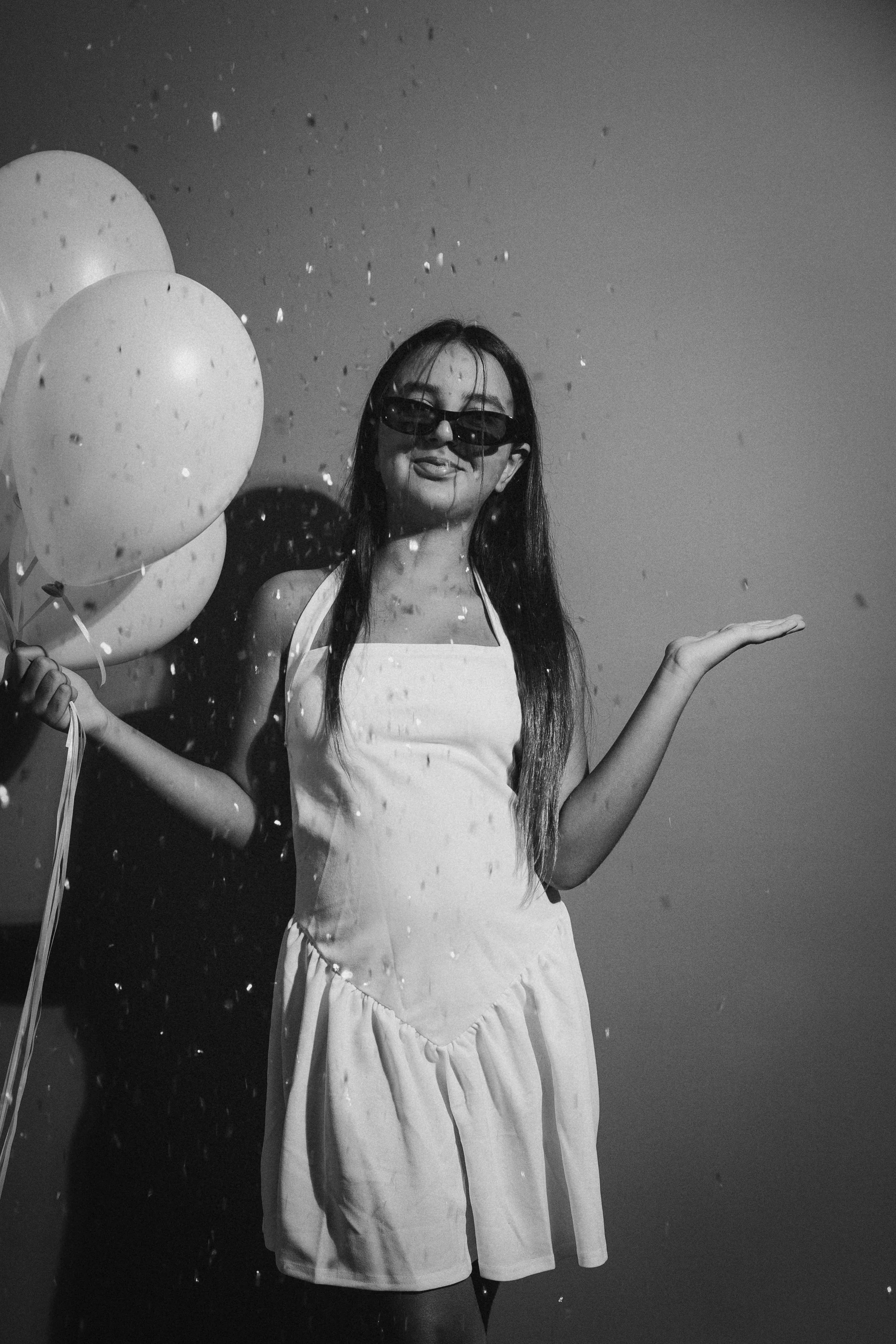 A woman with long hair wearing sunglasses and a white tank top, holding balloons and surrounded by falling confetti, posing outdoors against a cloudy sky.