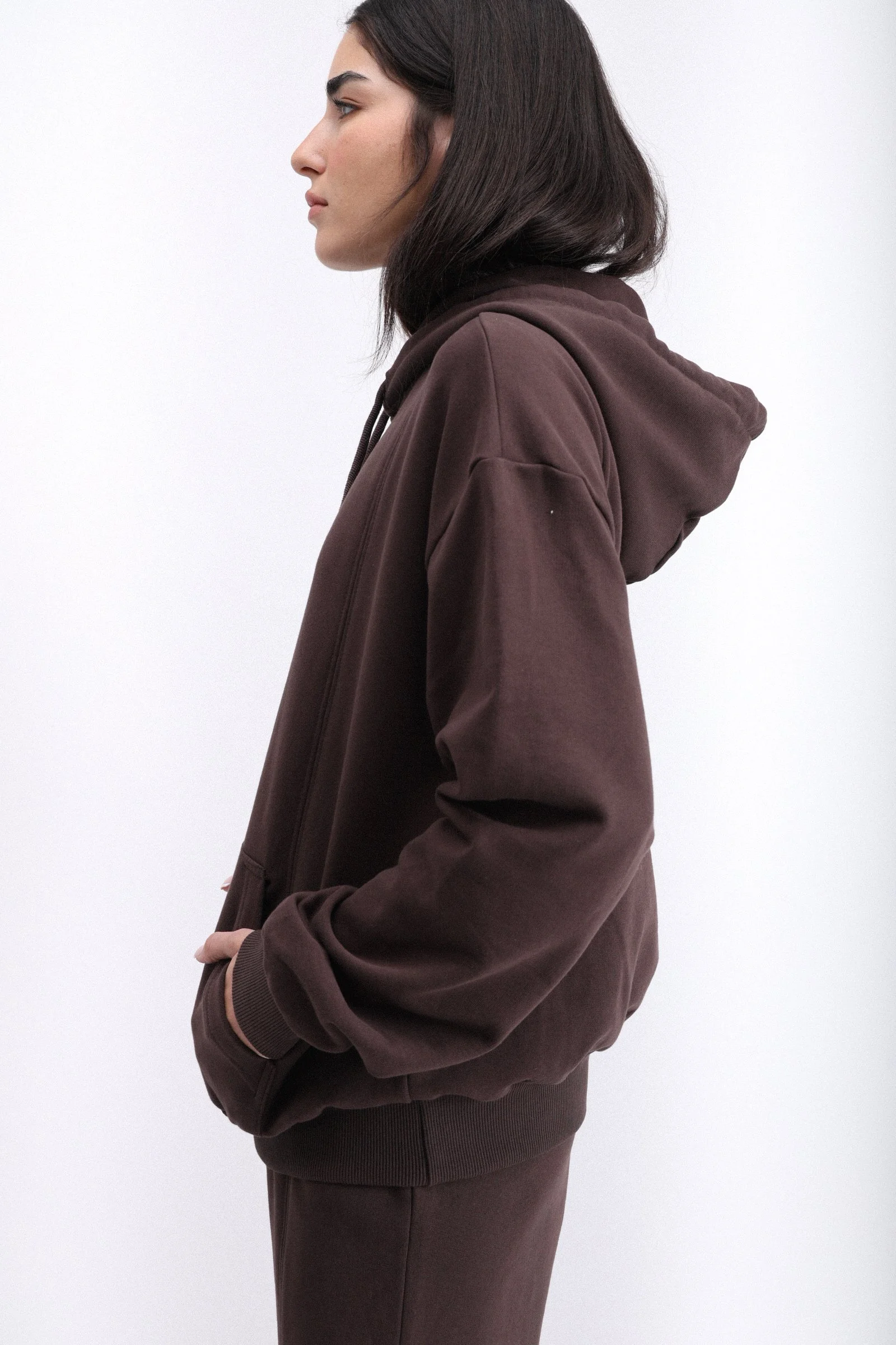 Side profile of a woman wearing a dark brown hoodie and matching pants, standing against a plain white background.