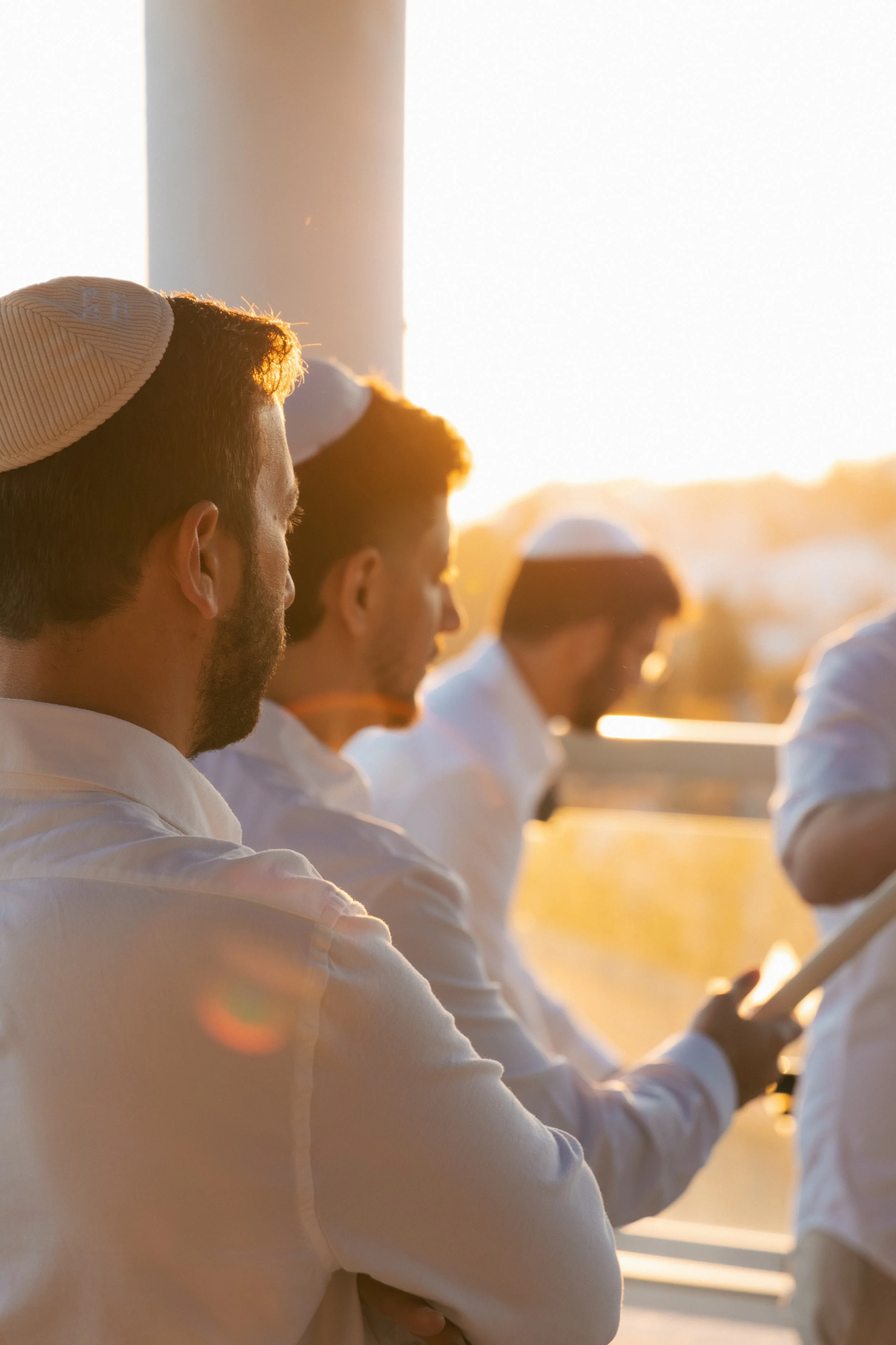 A group of men wearing white shirts and kippahs, standing outdoors at sunset, with a white structure and blurred background.