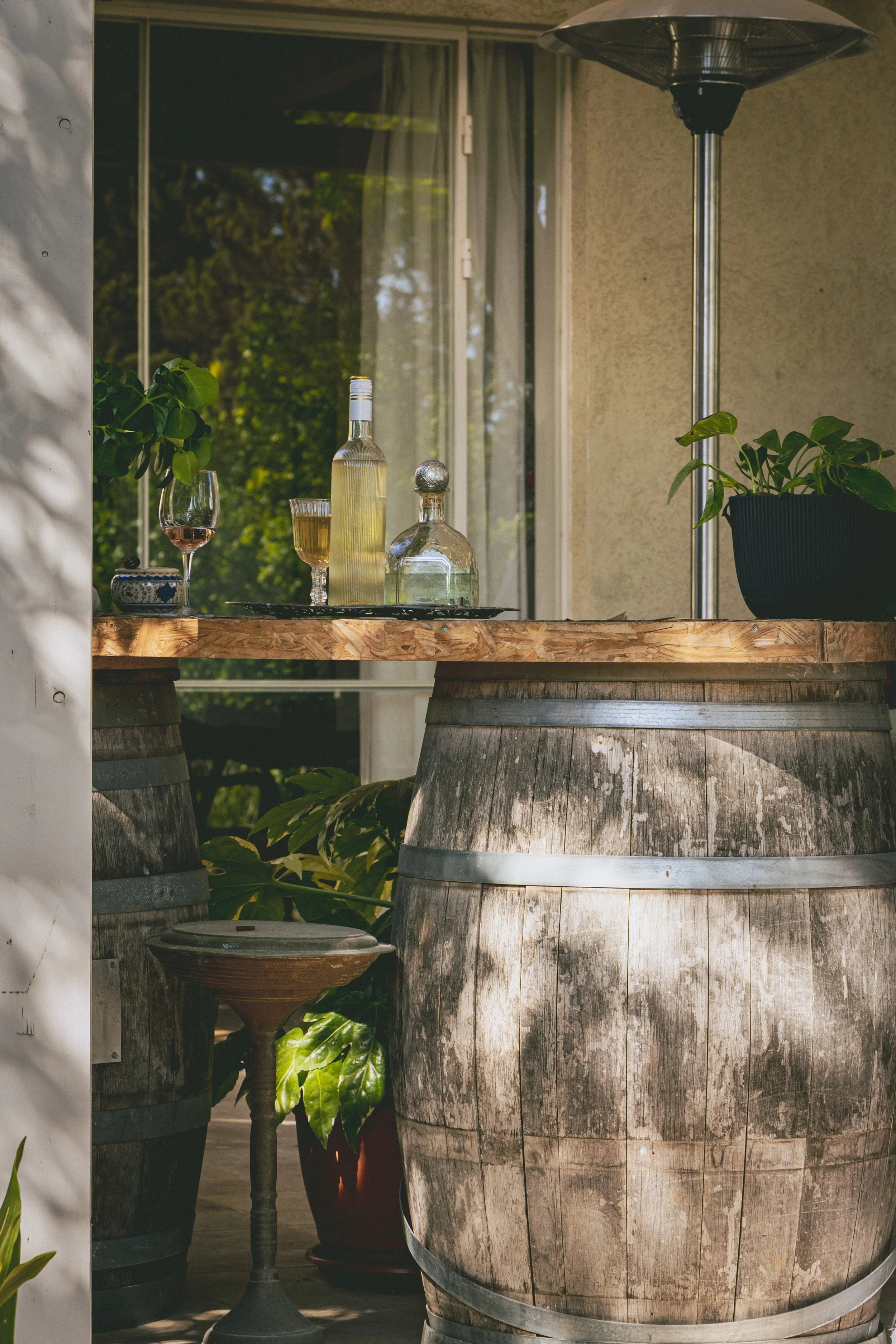 Outdoor scene showing two large wine barrels converted into tables with glass bottles, glasses of wine, and a potted plant on top. Background features a window and a patio heater.