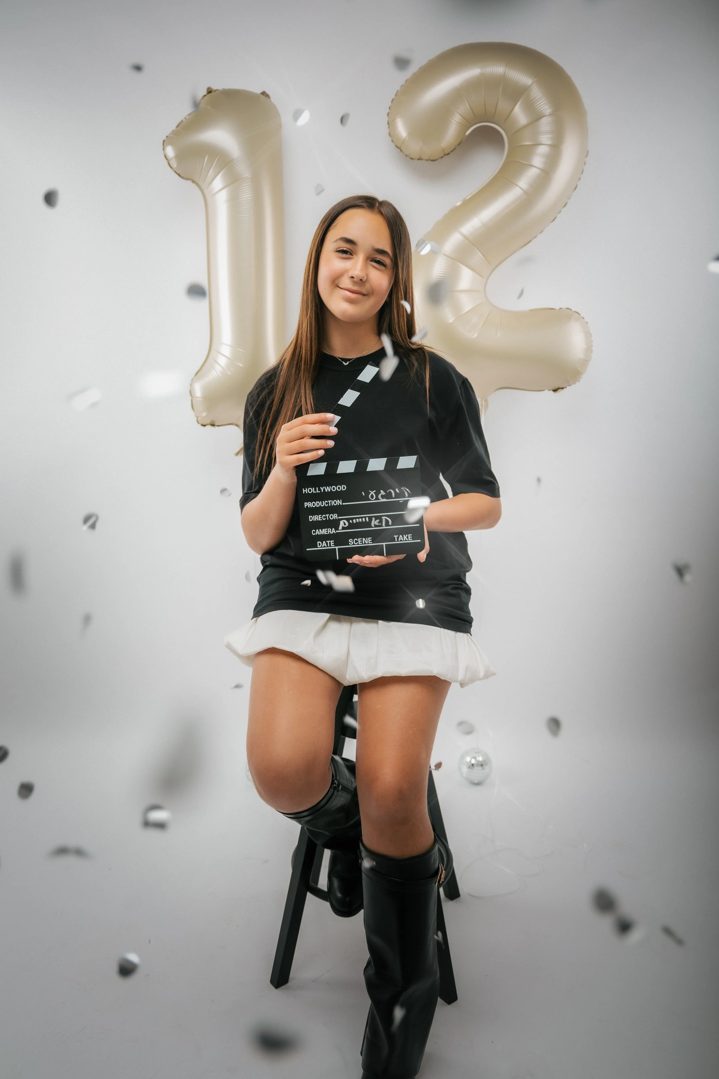 A young woman sits on a high stool, holding a clapperboard, with large gold balloons forming the number 12 behind her. She is smiling and surrounded by falling confetti in a celebratory setting.