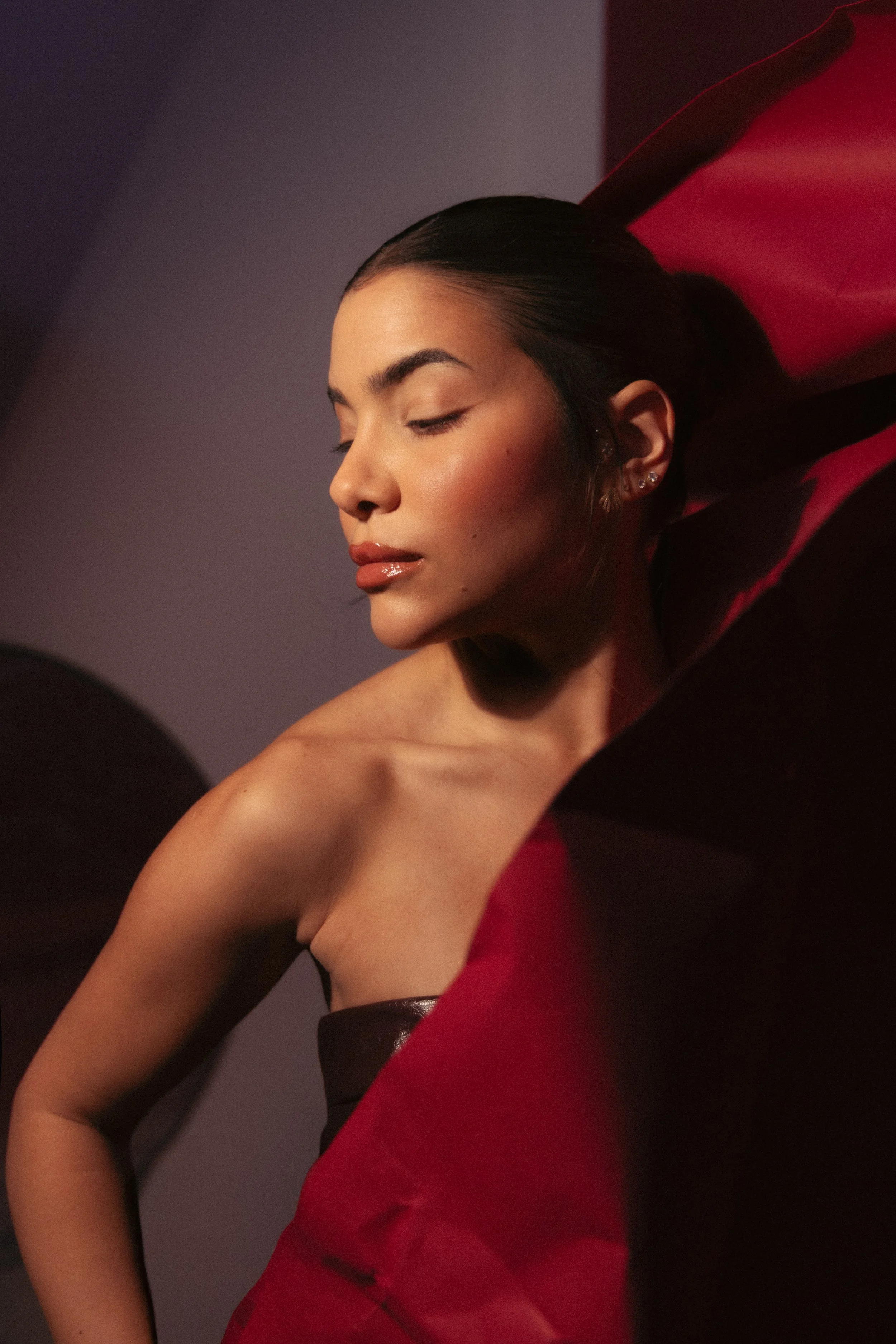 A woman with dark hair and earrings is leaning back with her eyes closed, wearing a red and black top, against a plain background.