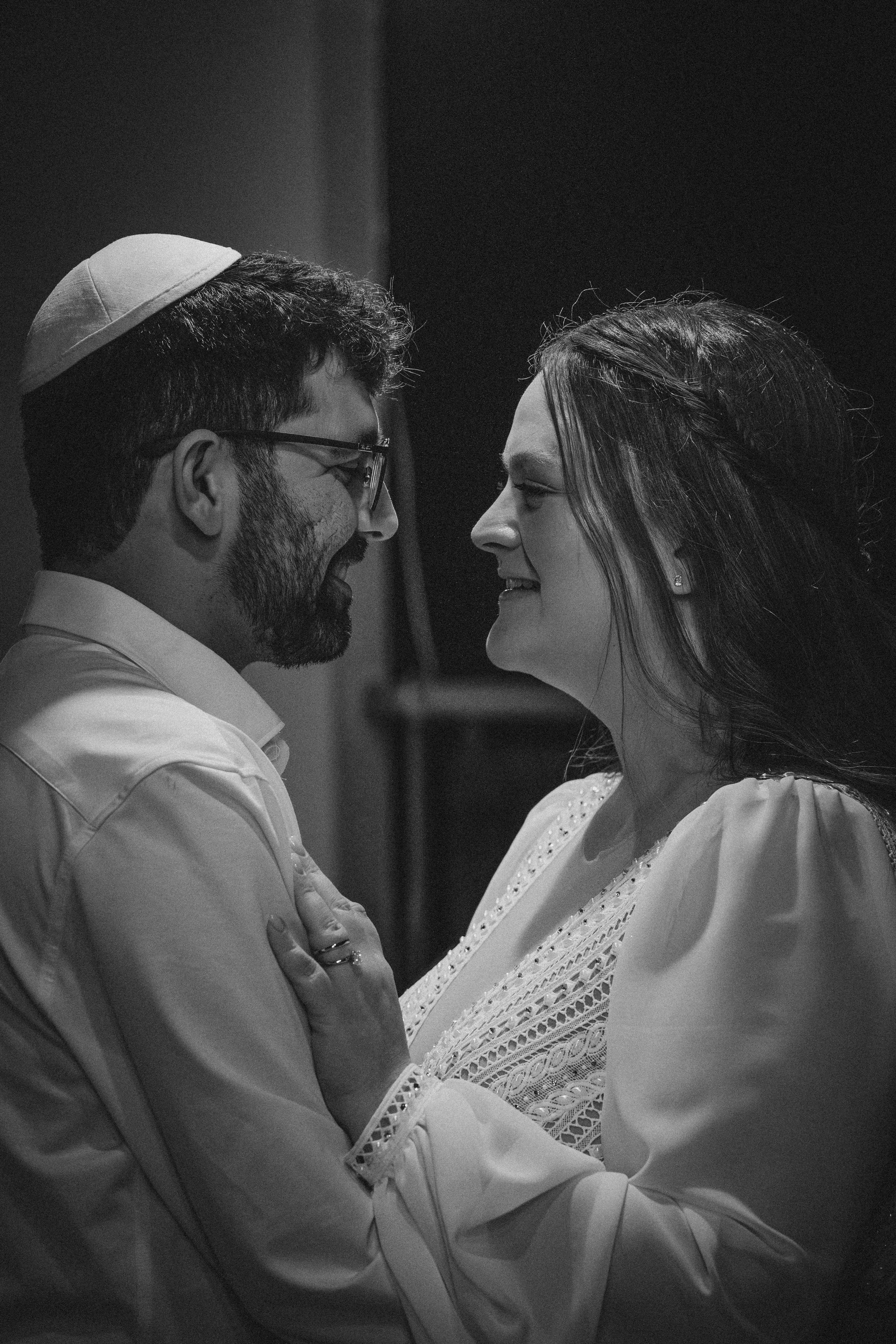 A black and white photo of a man and woman facing each other, smiling, with their foreheads touching. The man is wearing a kippah and glasses, and the woman is wearing earrings and a blouse with lace details.