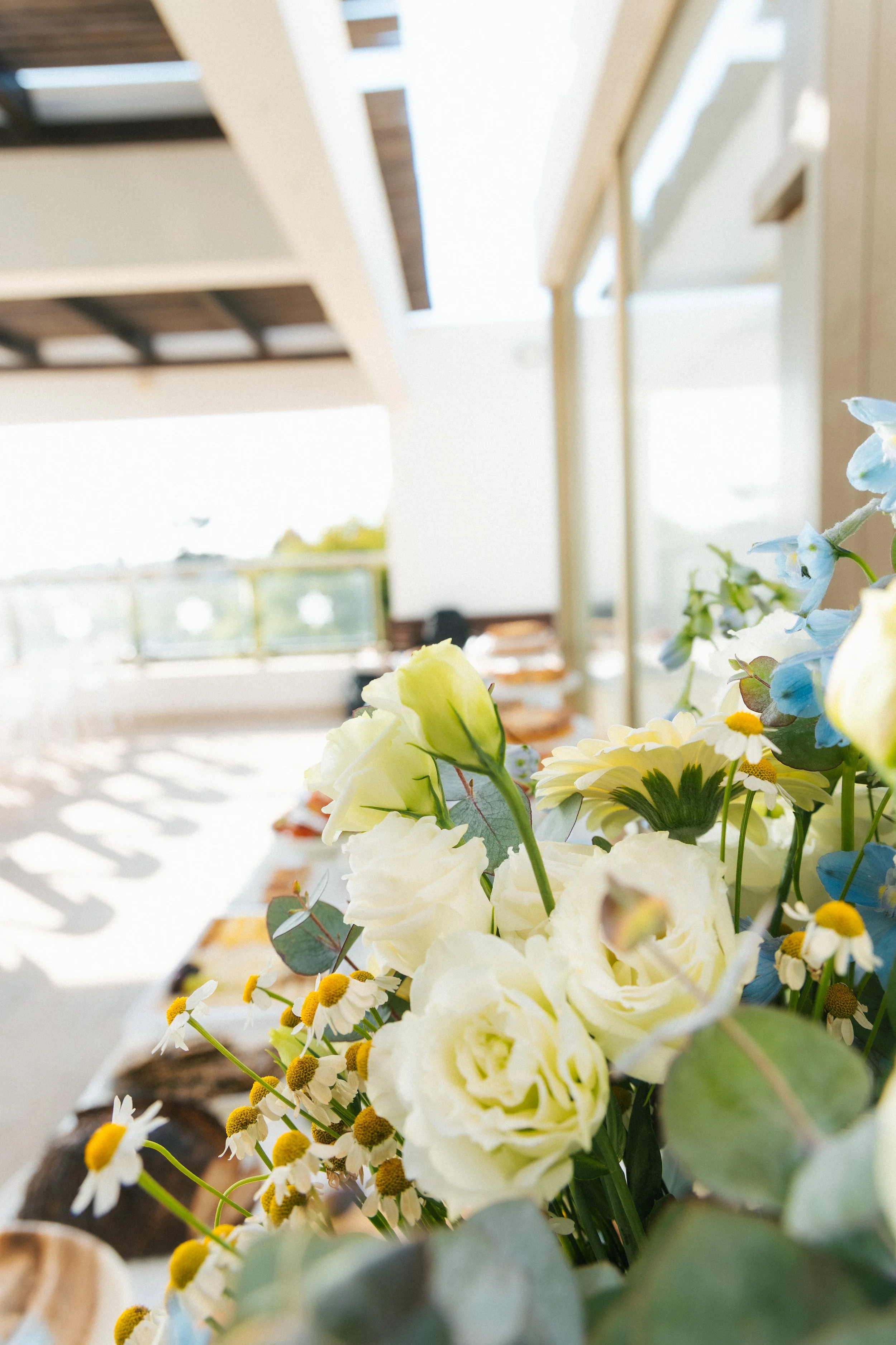 Close-up of a bouquet of white, yellow, and blue flowers with green leaves, in a well-lit room with large windows and a wooden ceiling.