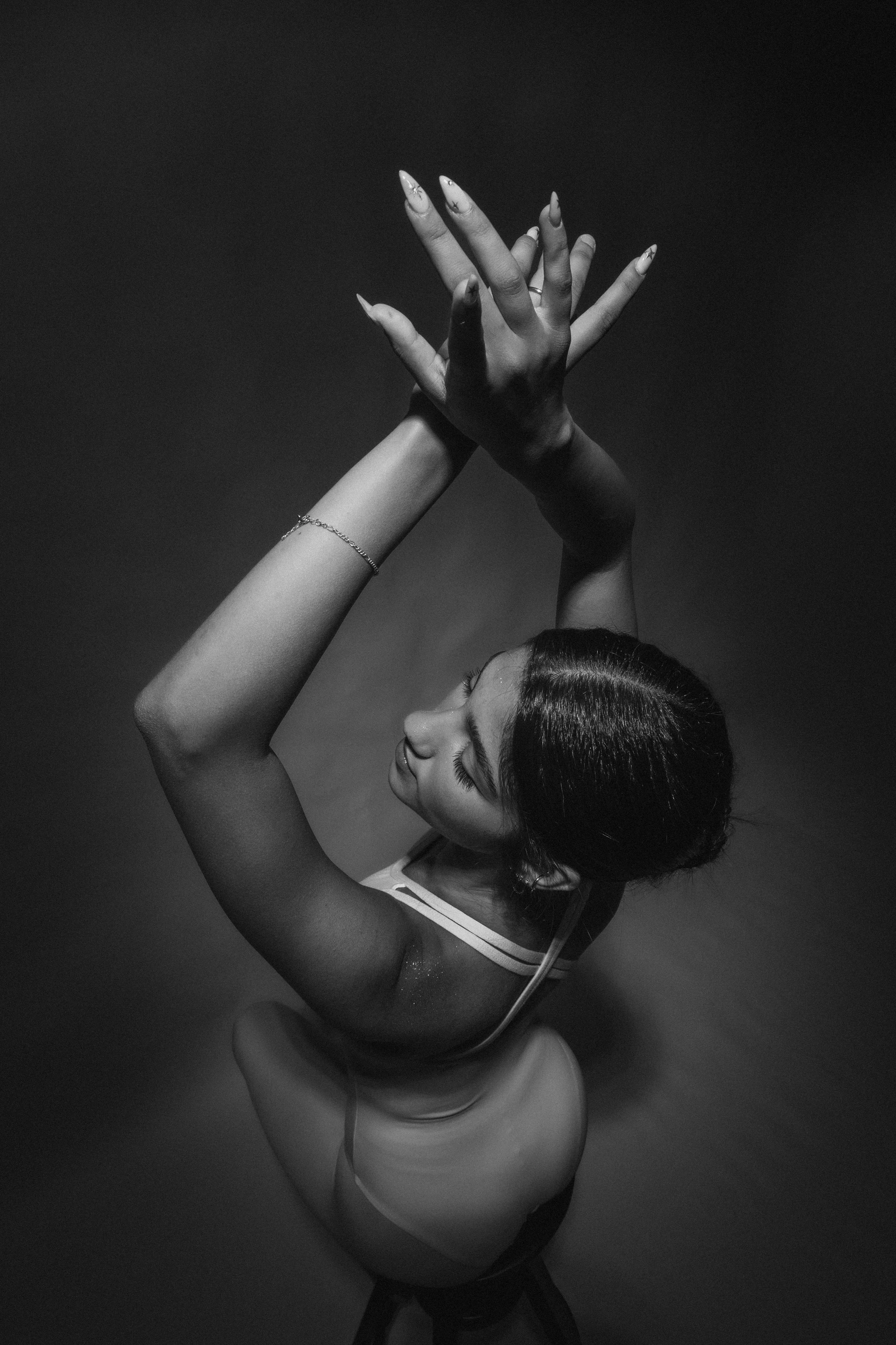A woman with dark hair balled up in a bun, wearing a sleeveless leotard, striking a dance pose with her arms raised and her fingers interlocked against a dark background.