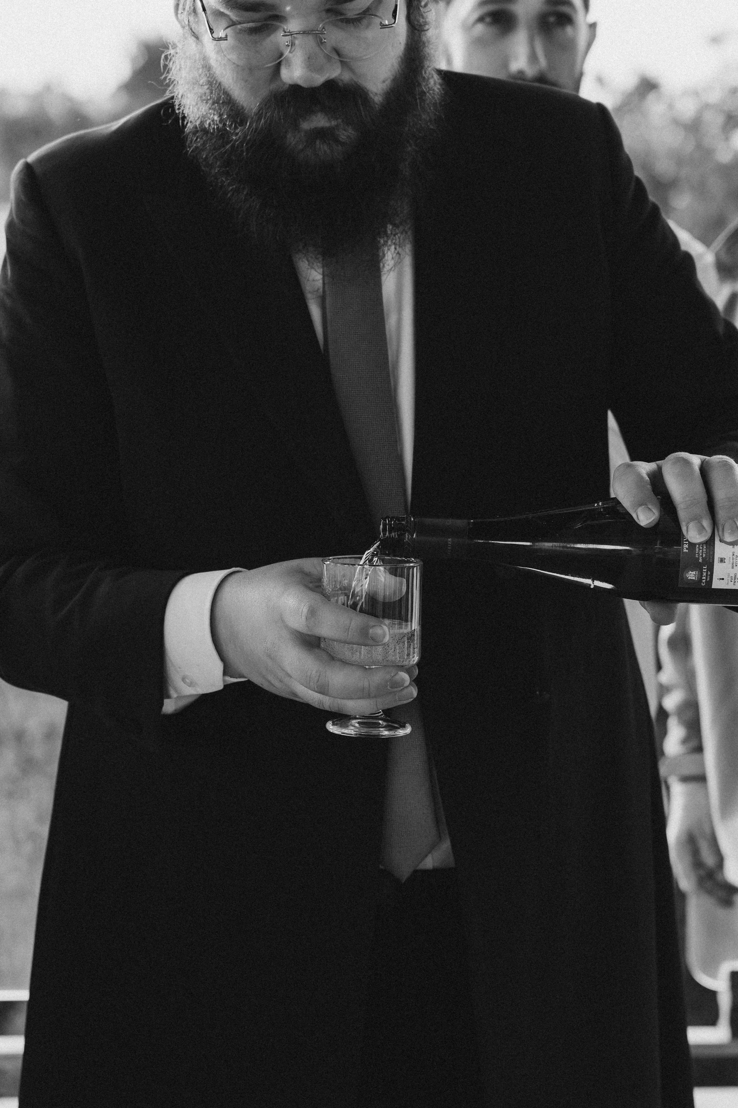Two men in formal attire, one with a beard and glasses pouring a beverage from a bottle into a glass, the other man observing in the background. The image is black and white.