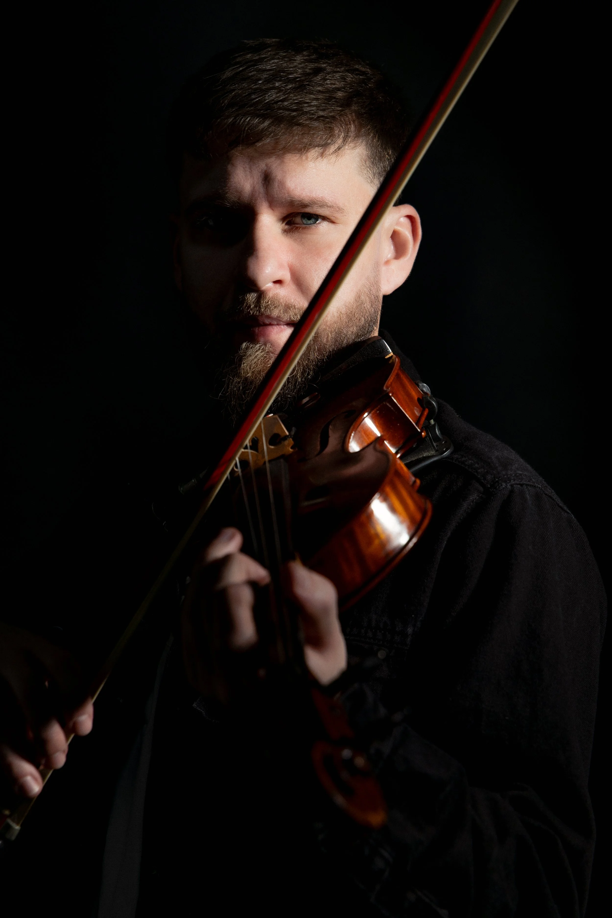 A man with a beard and short brown hair playing a violin against a dark background.