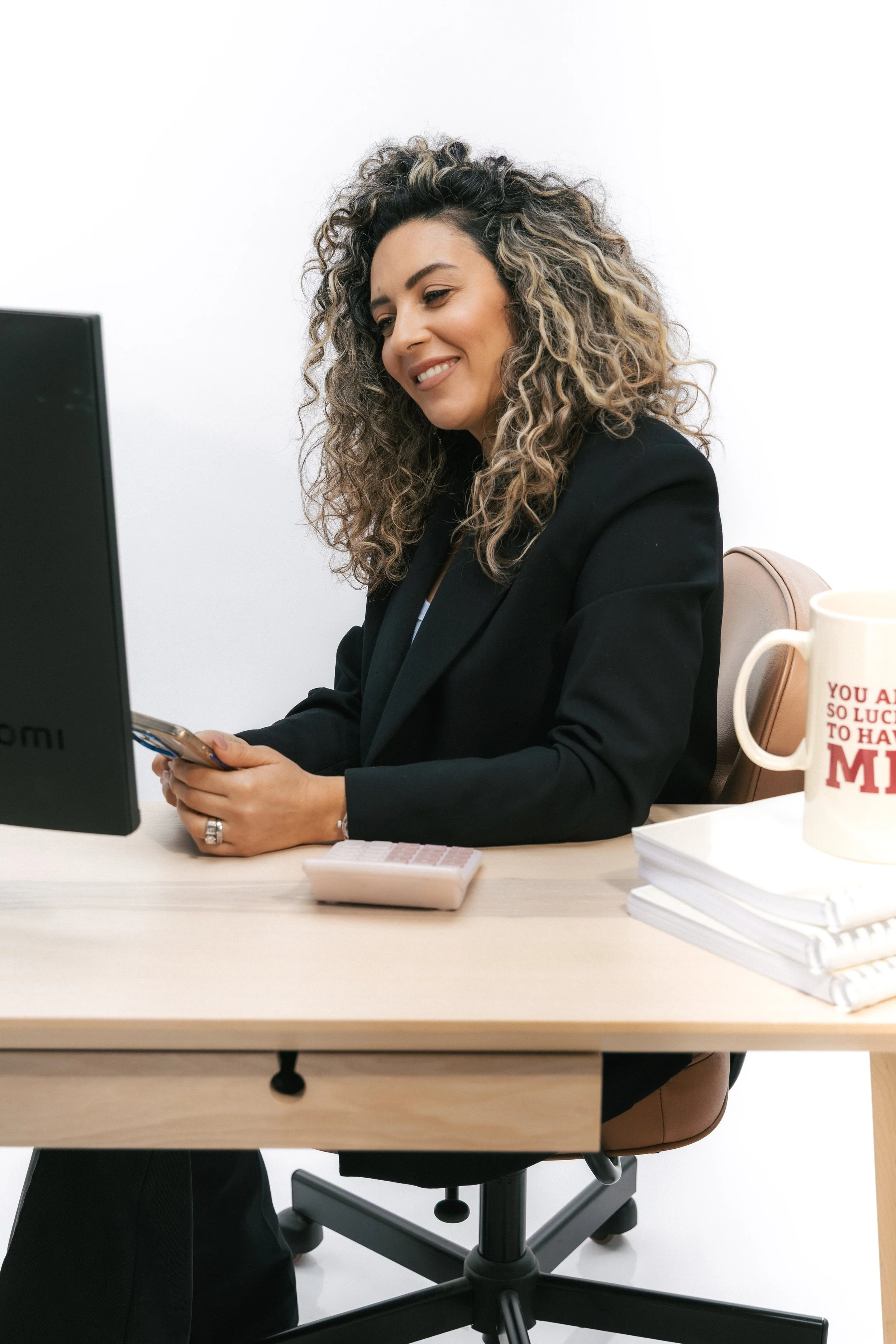 A woman with curly hair sitting at a desk, looking at her phone, with a computer and stacks of papers on the desk.