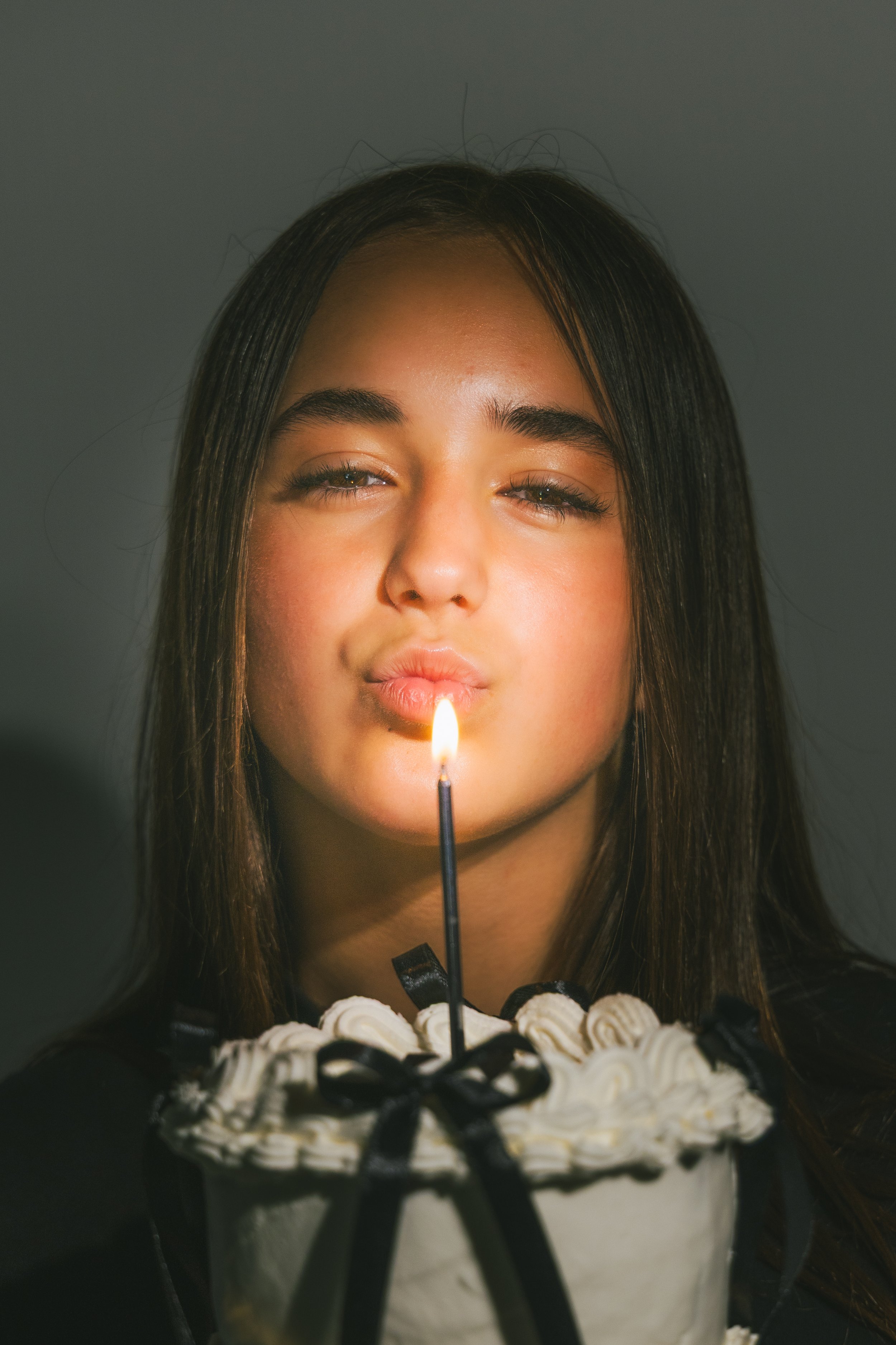 A woman with long brown hair is holding a lit birthday candle on top of a cake decorated with black ribbons and white frosting.