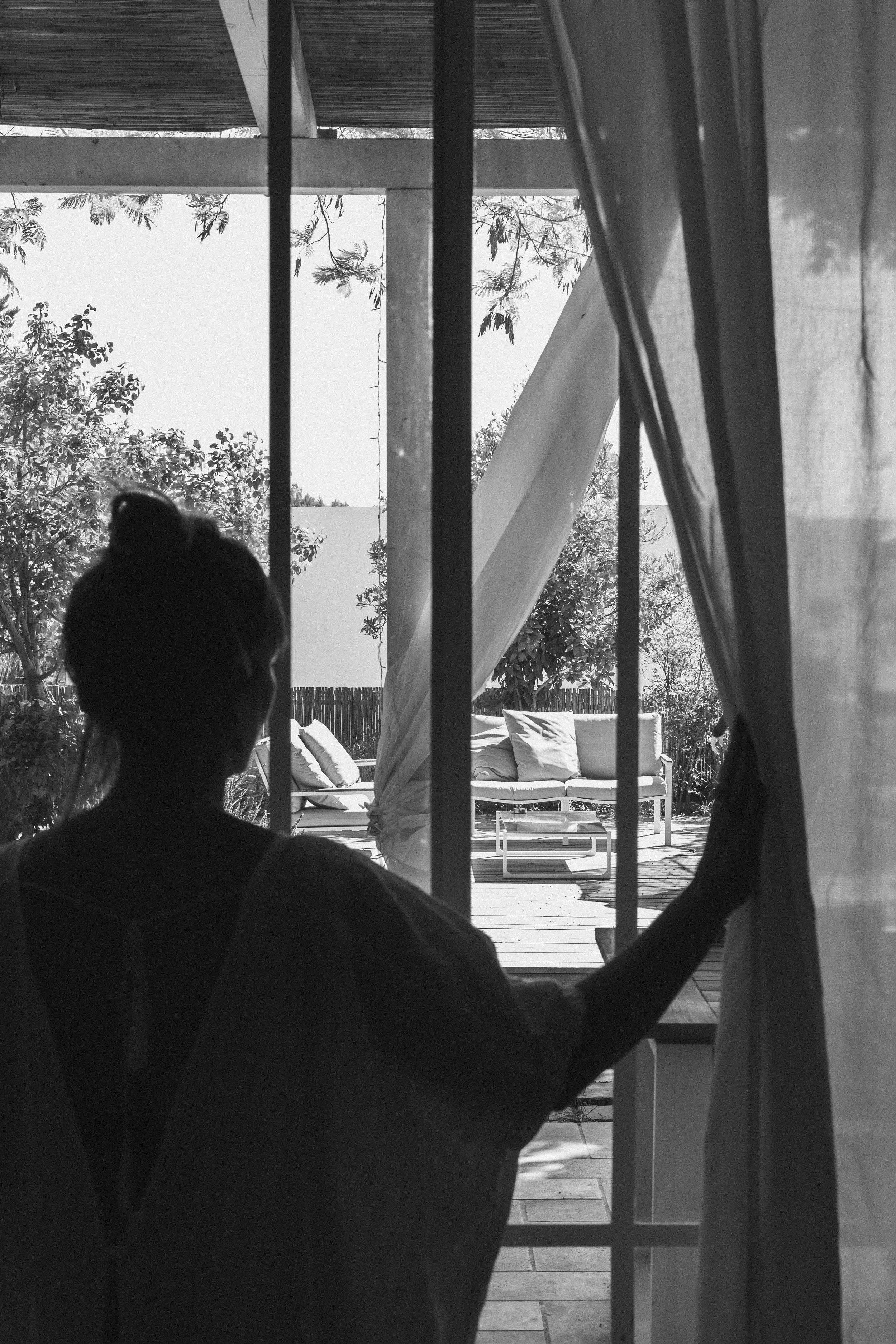 A woman is sitting at a table seen through a window with bars. Outside, there is a patio with a cushioned bench, trees, and a sunny sky.