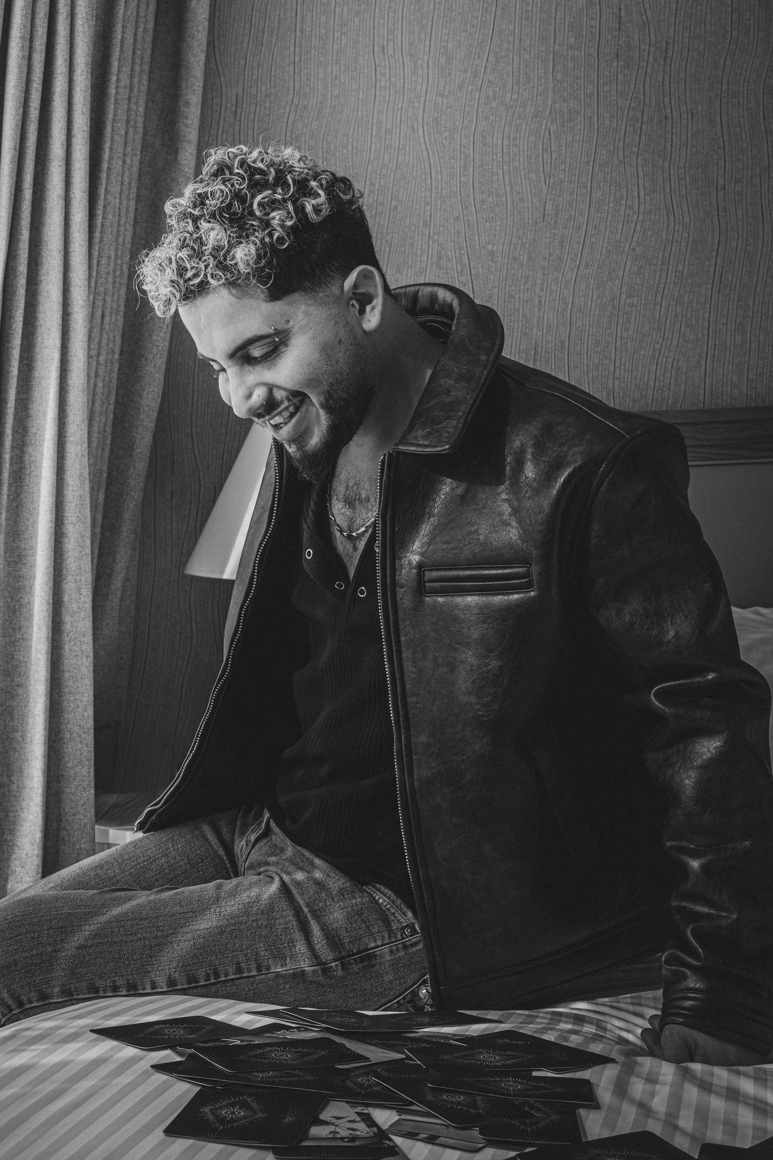 A man with curly hair and a beard, smiling, wearing a leather jacket, sitting on a bed with playing cards spread out on a checkered bedsheet, in a room with patterned wallpaper and a curtain.