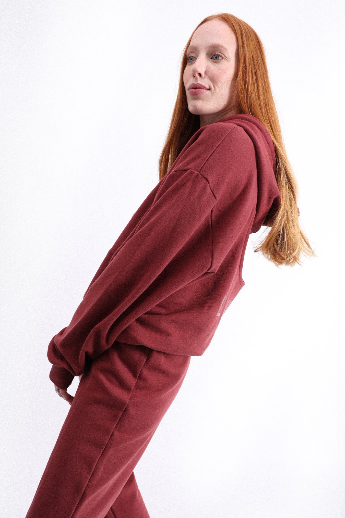 A young woman with long red hair and fair skin wearing a matching maroon hoodie and sweatpants, standing against a plain white background.