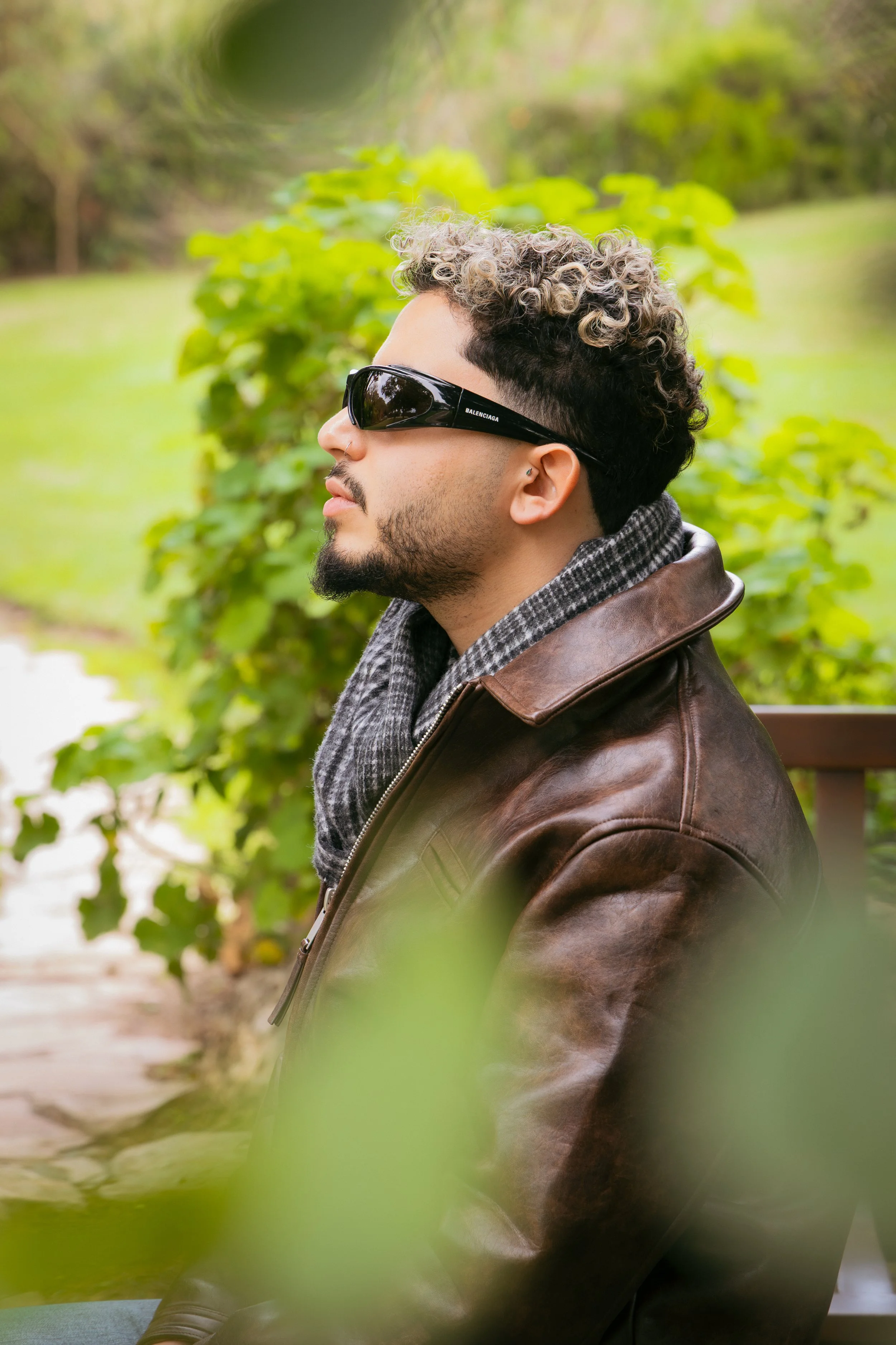 A man with curly blond and black hair, wearing sunglasses, a nose ring, and a scarf, sitting outdoors on a park bench surrounded by green foliage.