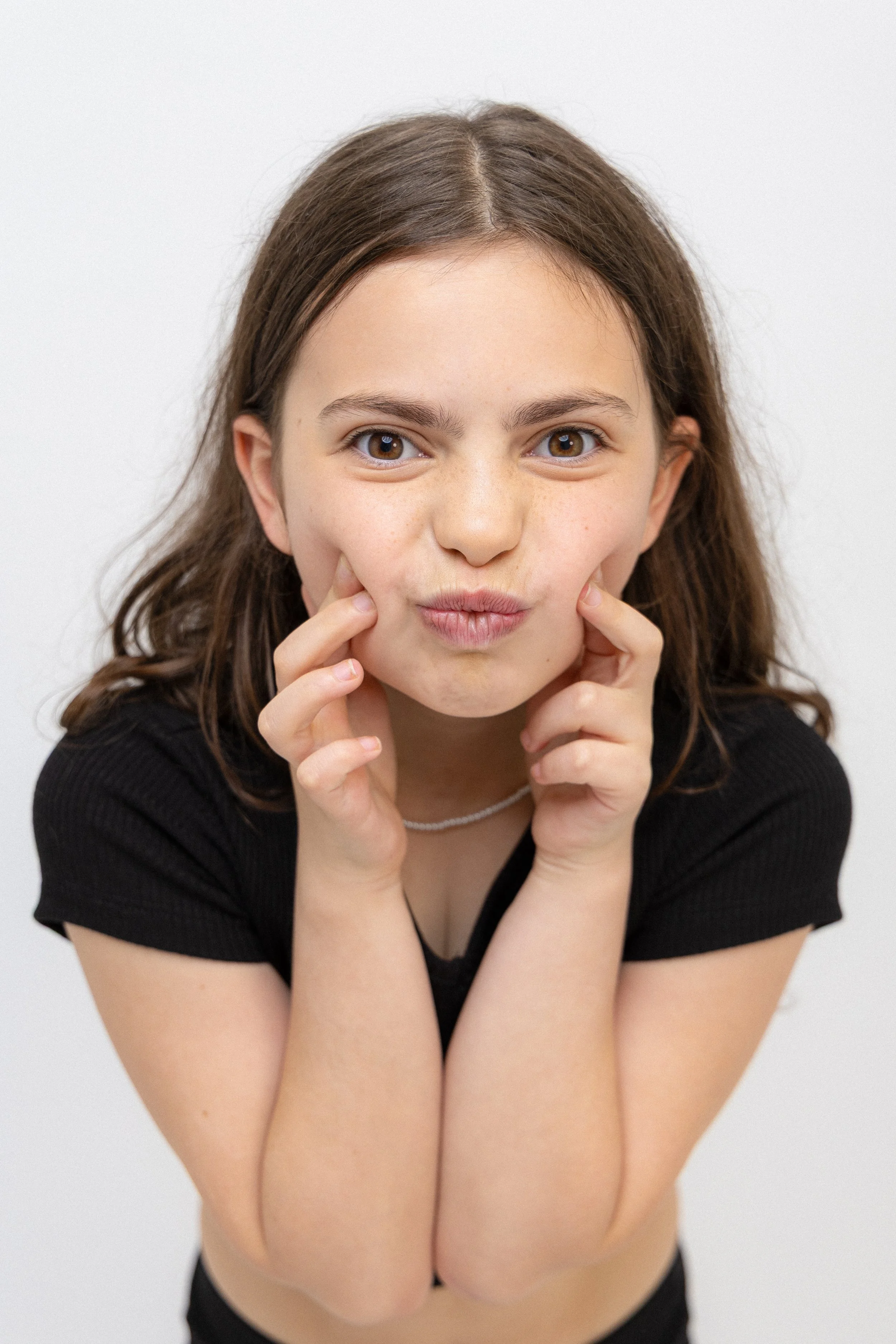 A young girl with brown hair and brown eyes wears a black top, touching her cheeks with her fingers, looking directly at the camera against a white background.