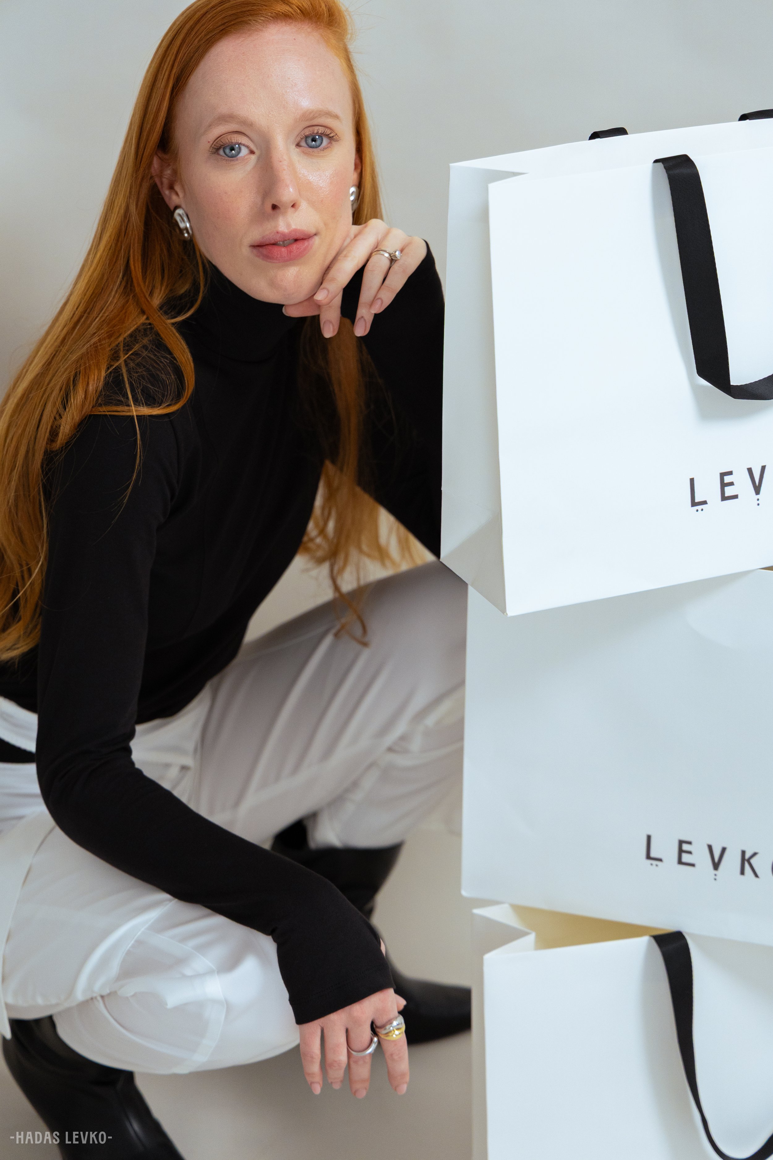 A woman with long red hair wearing a black turtleneck and white pants, sitting next to white shopping bags with black handles that have the brand name 'LEVK' on them.