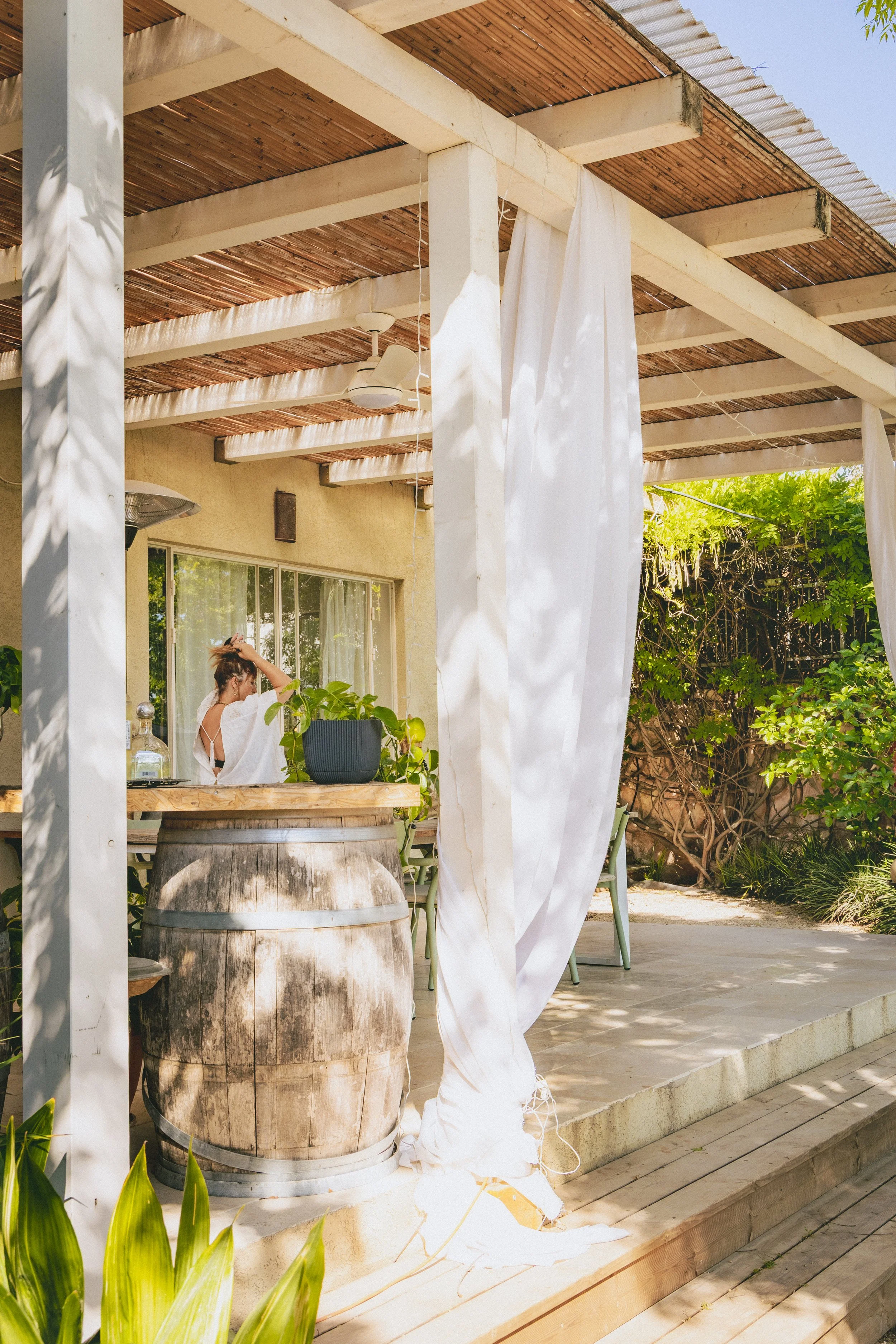 A woman in white dress standing outside on a patio, next to a large wooden barrel with plants on top, under a pergola with white curtains and wood ceiling, surrounded by greenery.