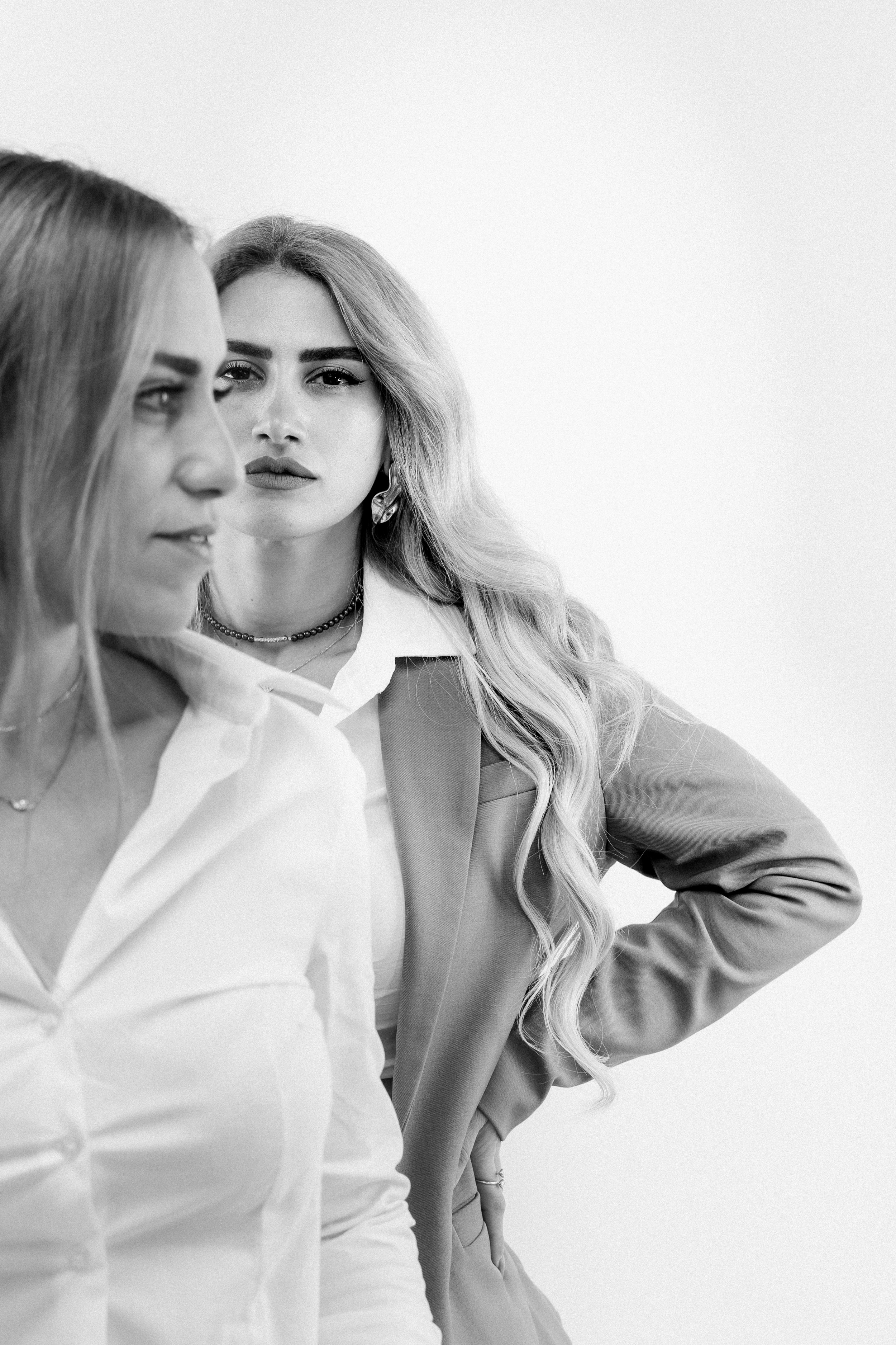 Black and white photo of two women, one in the foreground with long hair and a collared shirt, partially visible, and the other in the background with wavy hair, wearing a blazer and earrings, looking directly at the camera.