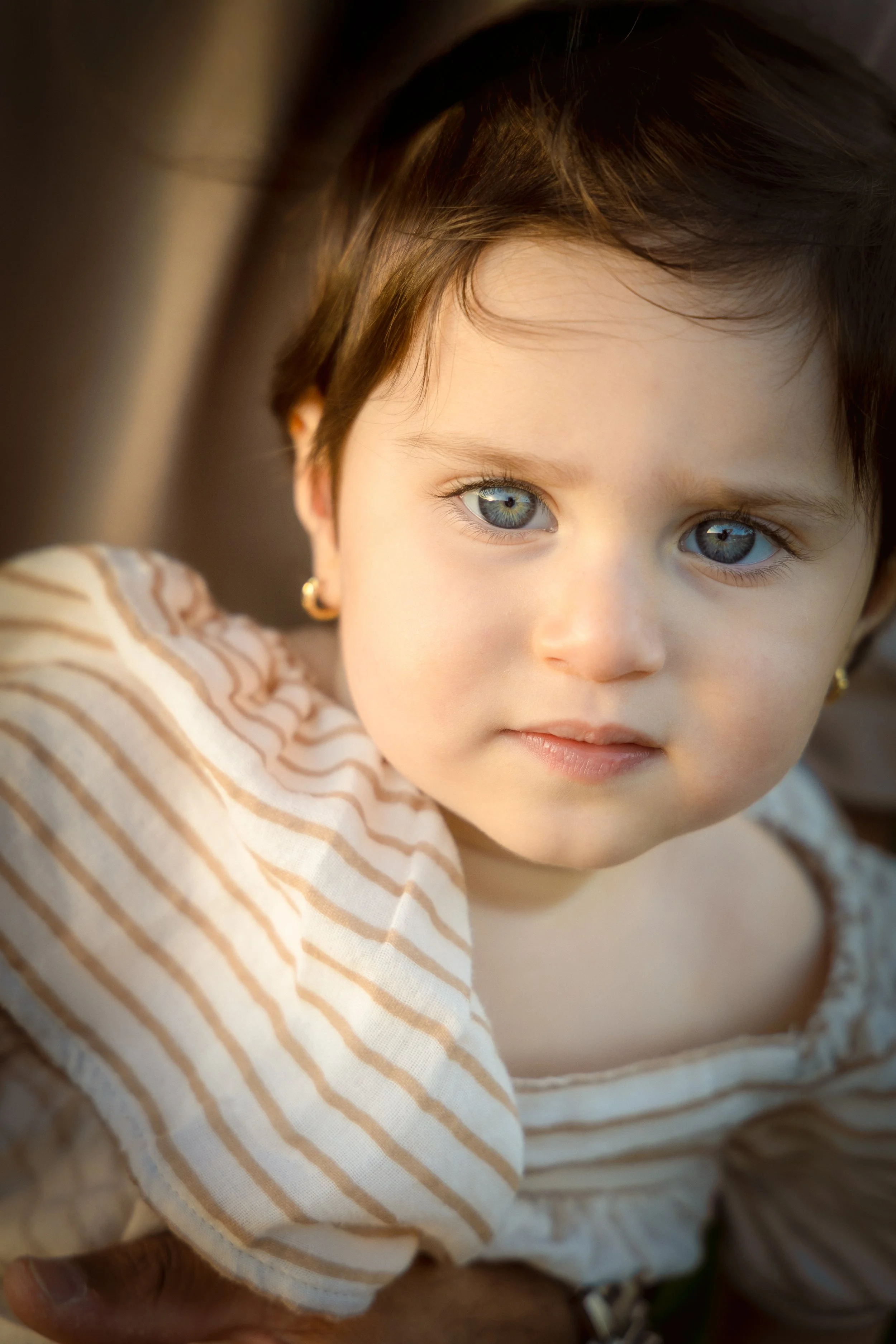 Close-up of a young girl with blue eyes, dark hair, wearing gold earrings and a cream-colored shirt with brown stripes, being held or supported by someone with darker skin.