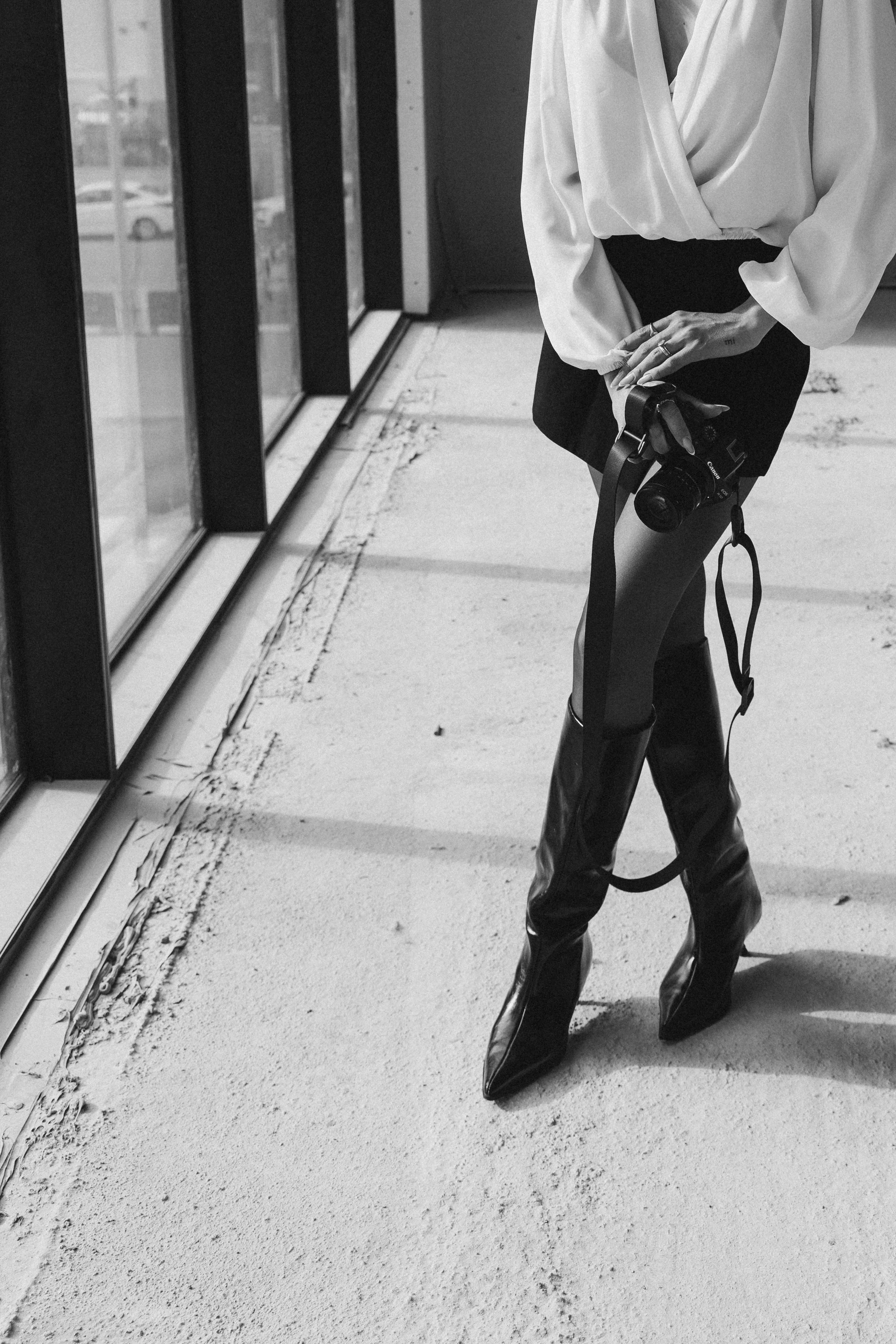 A woman is standing indoors near a glass wall, holding a camera. She is dressed in a loose blouse and shiny knee-high boots, with her hands clasped and a tattoo visible on her wrist.