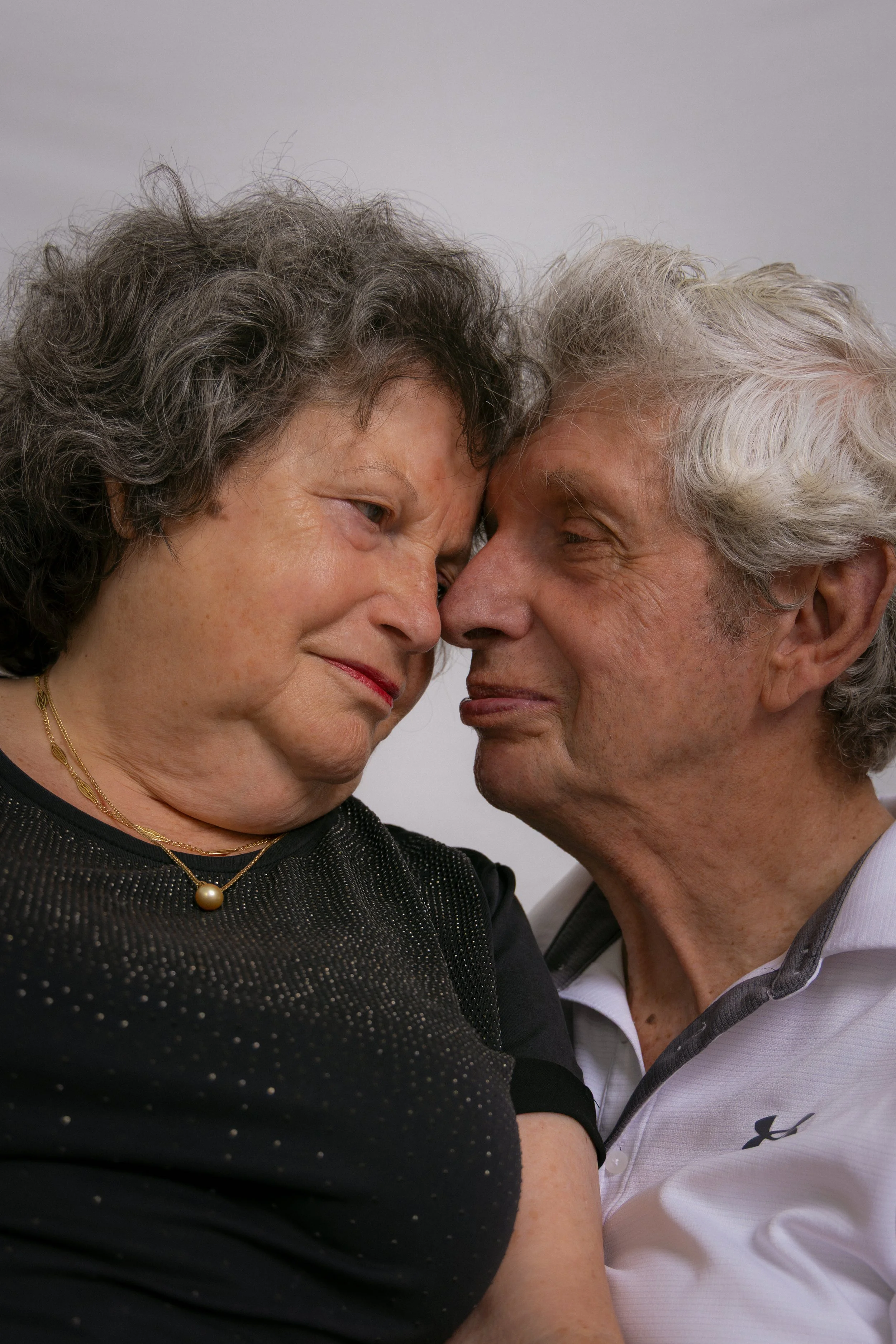 An elderly man and woman are touching foreheads, with eyes closed, creating an intimate and tender moment against a plain background.