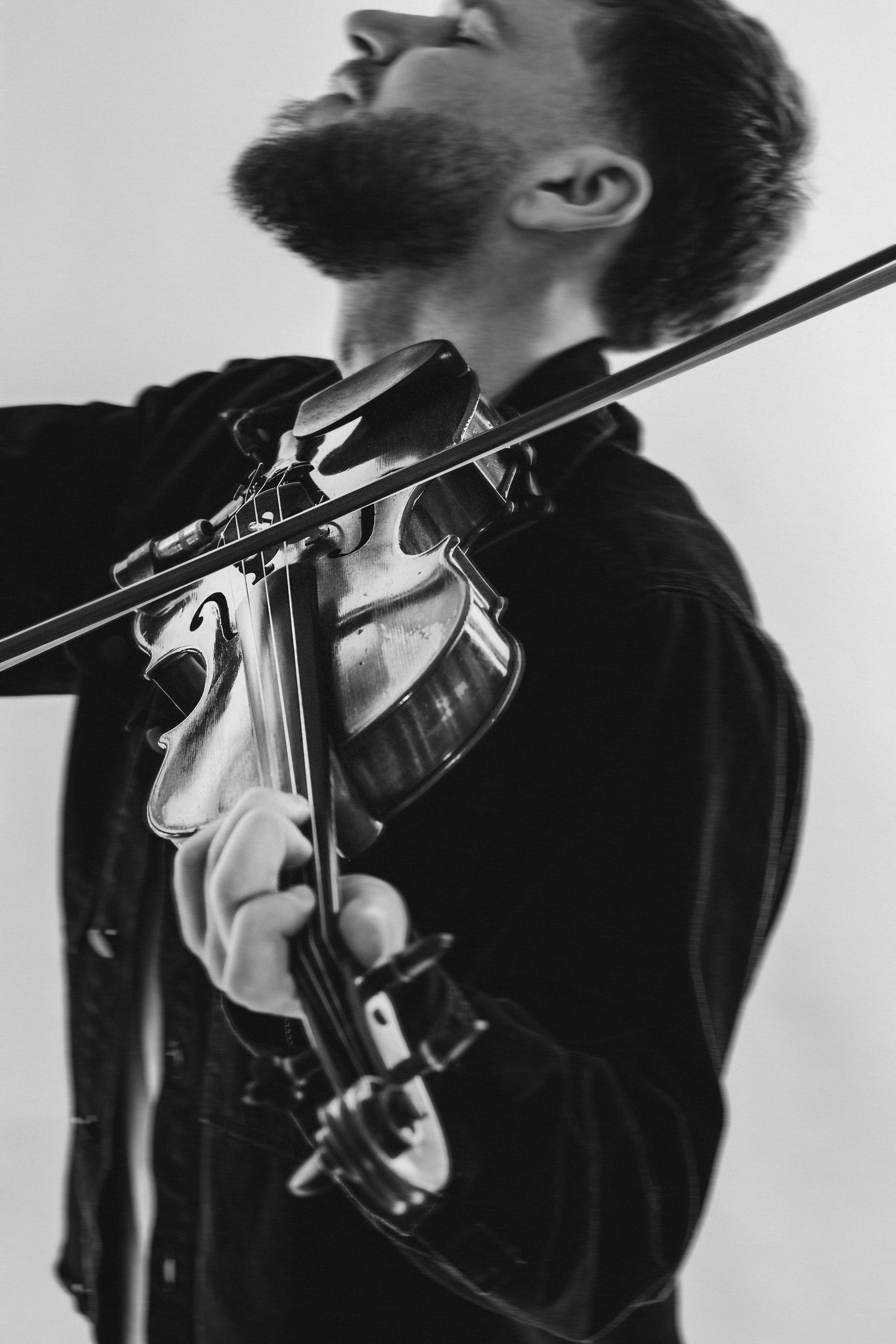 A black and white photo of a man playing the violin, viewed from a low angle showing his face in profile and his hand holding the violin's neck.