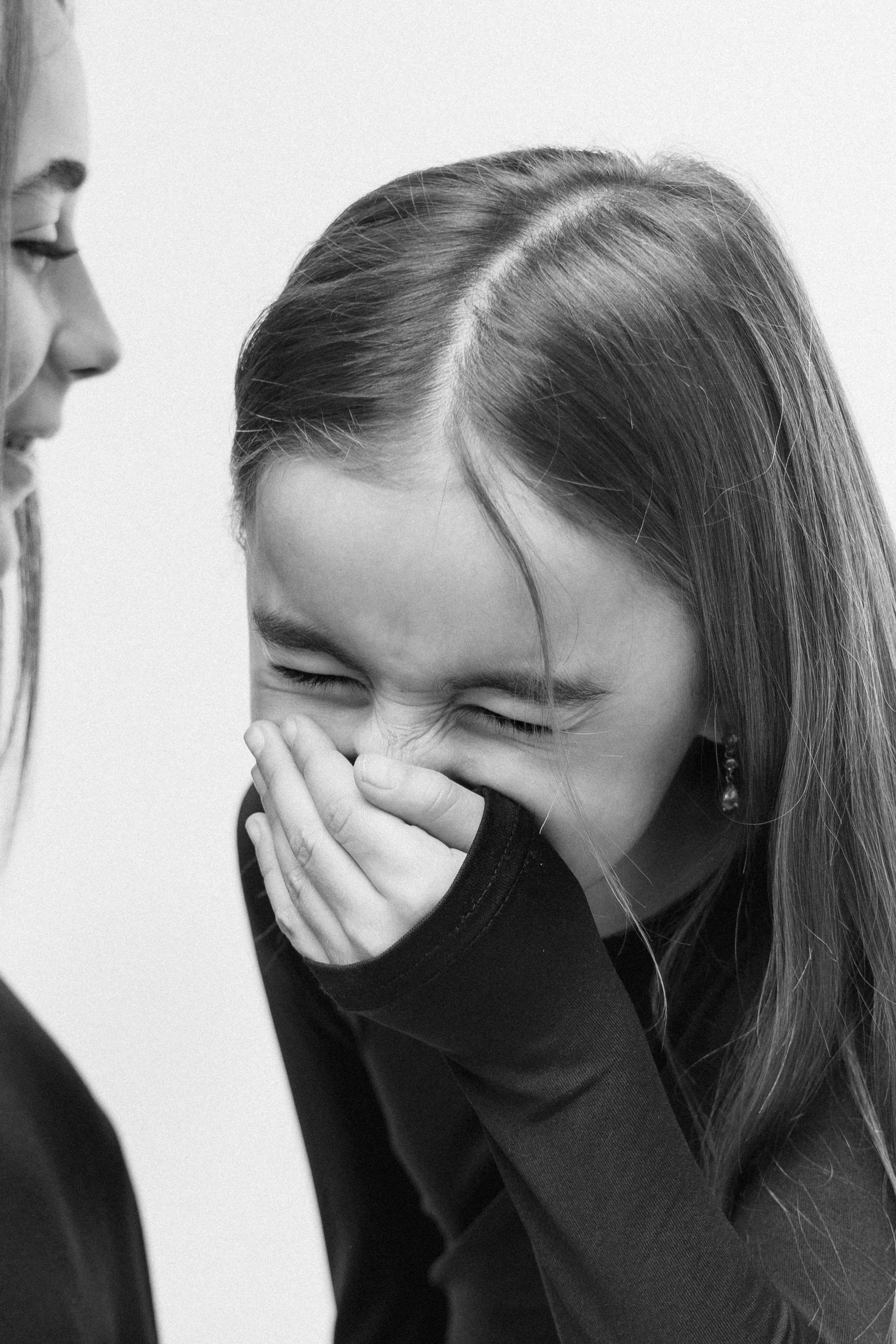 A young girl is laughing and covering her mouth with her hands while an adult woman, possibly her mother, smiles at her. The image is in black and white, capturing a moment of joy and connection.