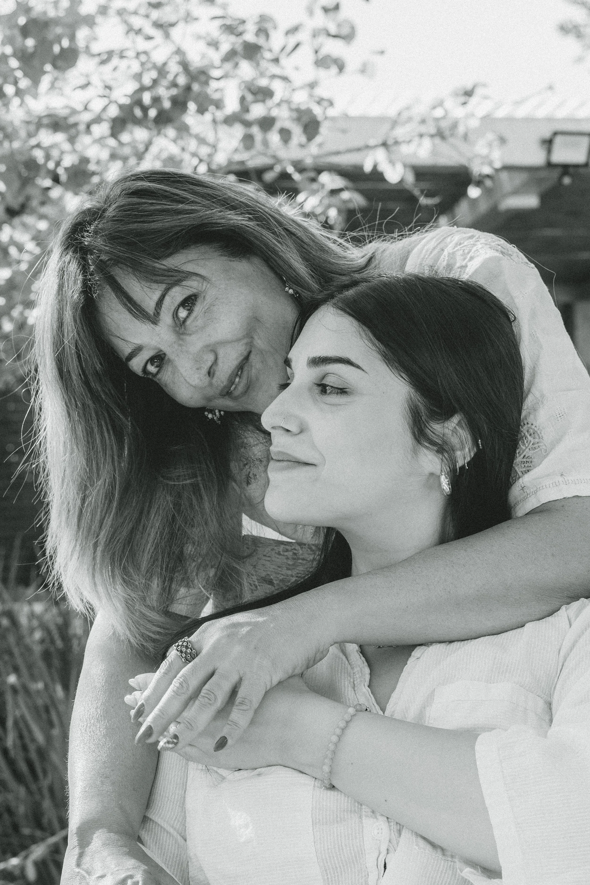 A black and white photo of a woman with dark hair and another woman with lighter hair. The woman with lighter hair is resting her head on the woman with dark hair, smiling. They appear to be outdoors with trees and a building in the background.