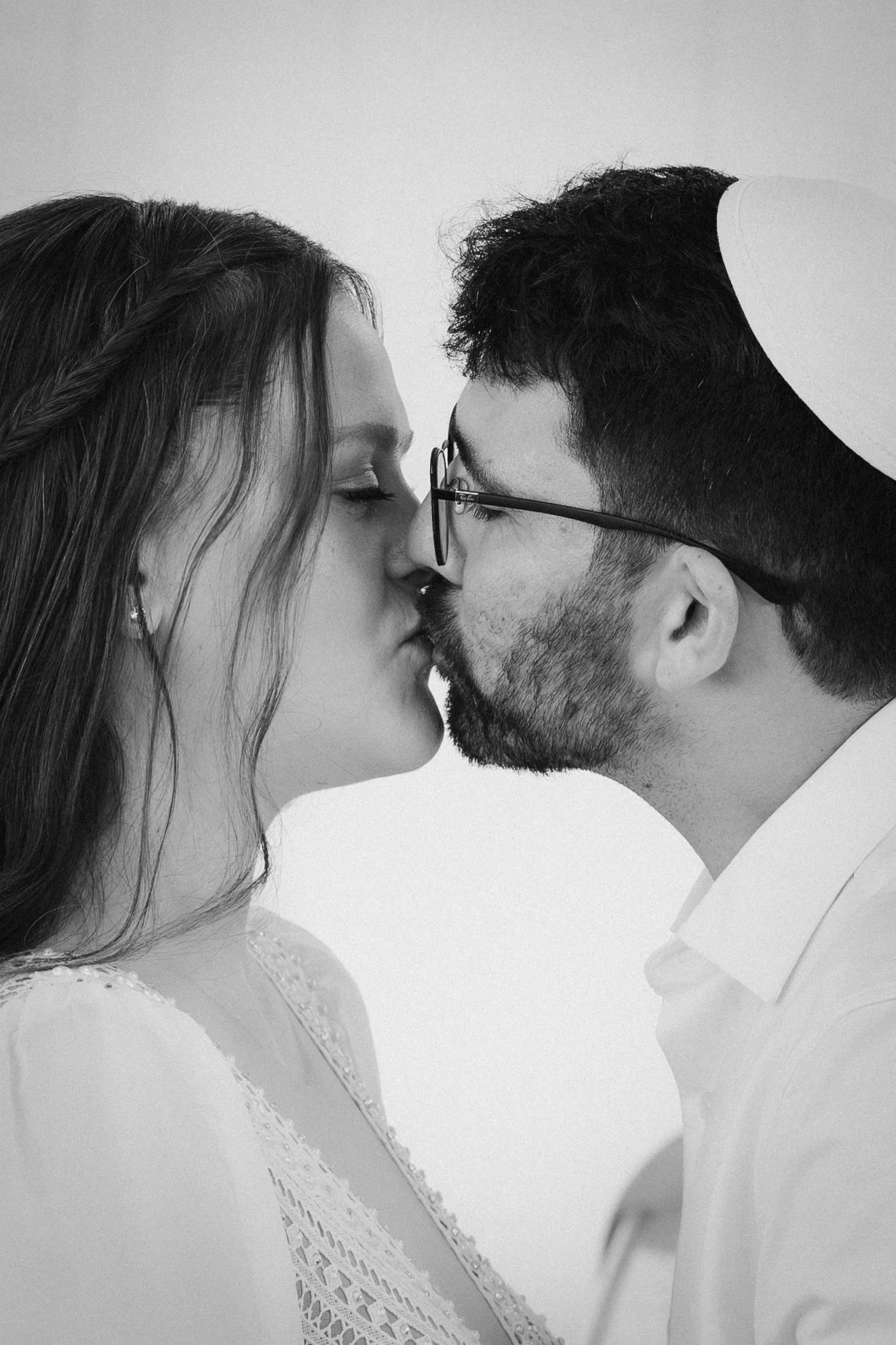 Black and white photo of a couple with foreheads touching, eyes closed, about to kiss. The woman has long hair in a braid and wears a lace dress. The man wears glasses and a white shirt.