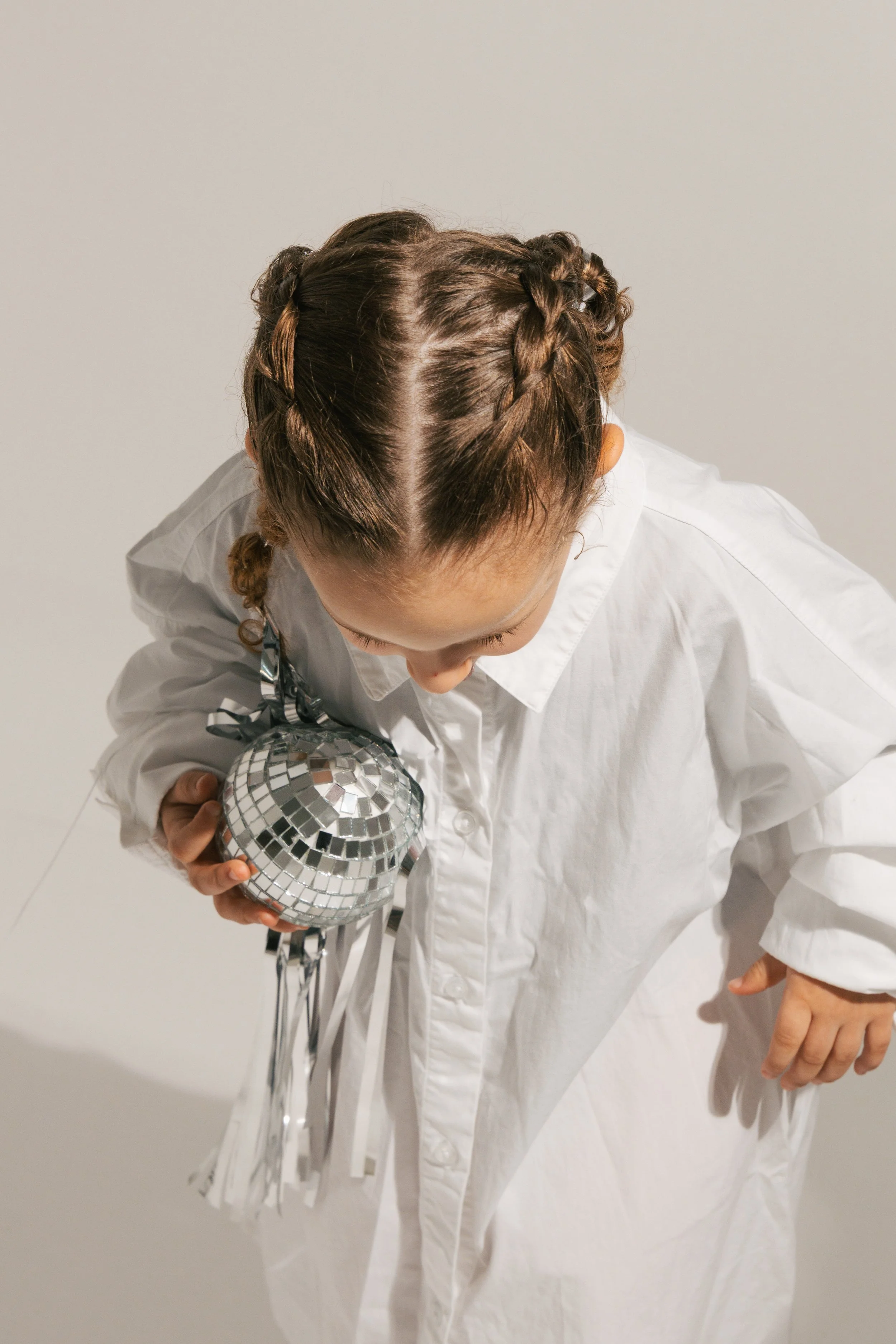 Person with braided hair and white shirt holding a small disco ball, standing against a plain light-colored wall.