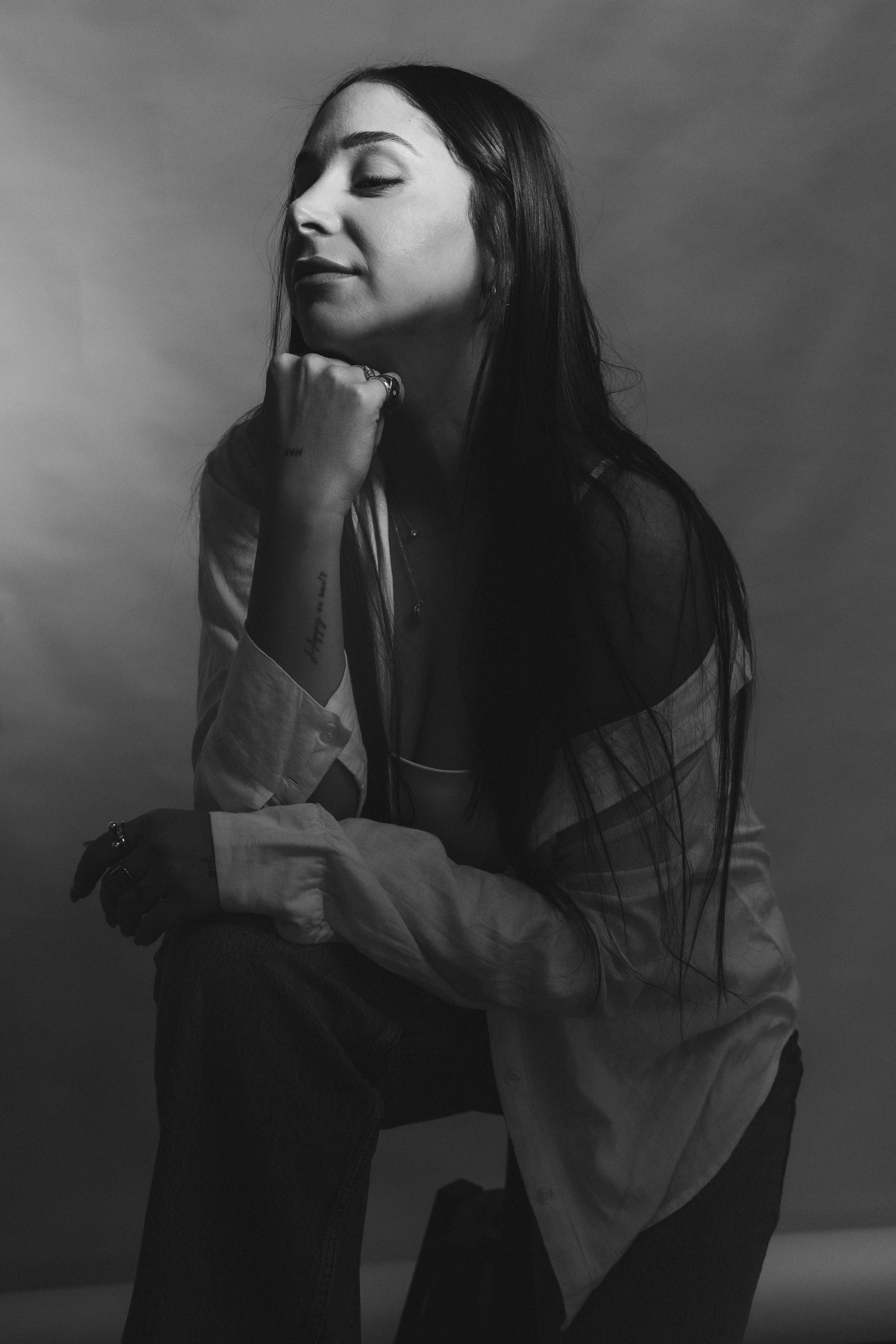 Black and white portrait of a woman with long dark hair, closed eyes, resting chin on her hand, wearing rings, a necklace, a light-colored shirt, and dark pants, against a plain background.