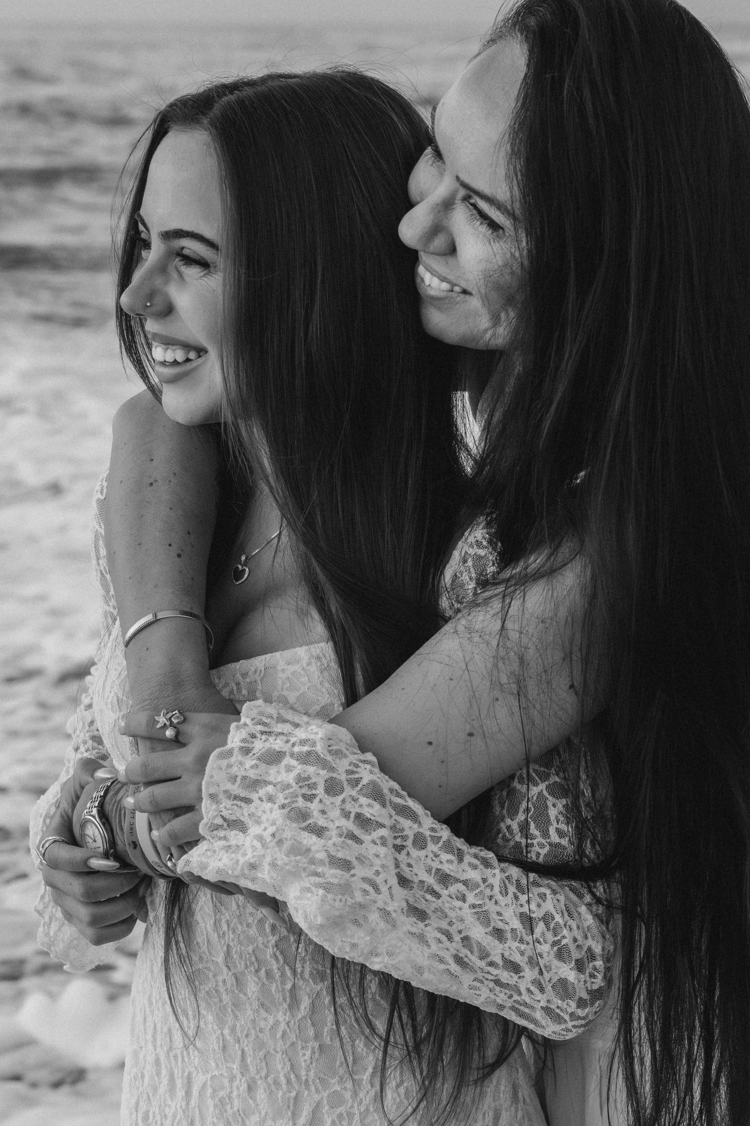 Two women with long dark hair smiling and hugging on a beach, wearing lace dresses, in black and white.