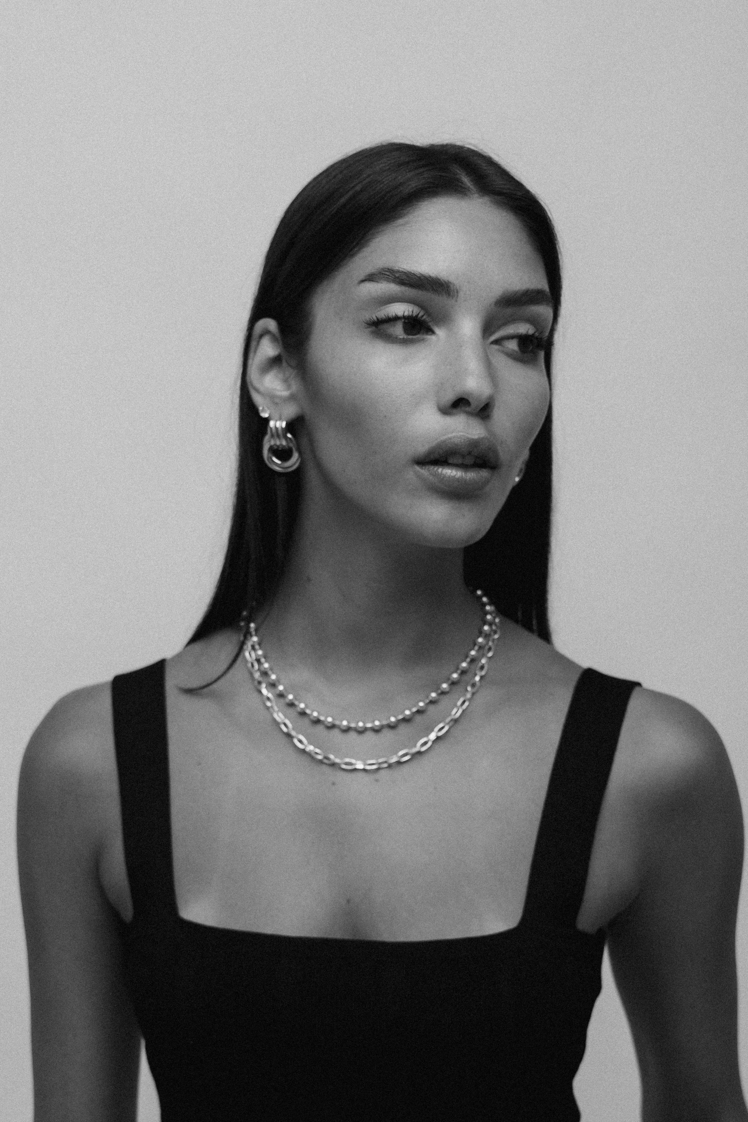 A black and white portrait of a woman with long dark hair, wearing layered necklaces, hoop earrings, and a sleeveless black top.editorial photoshoot. fashion photography. צילומי אופנה, צילומי תדמית, דוגמנית, שחור לבן, מ