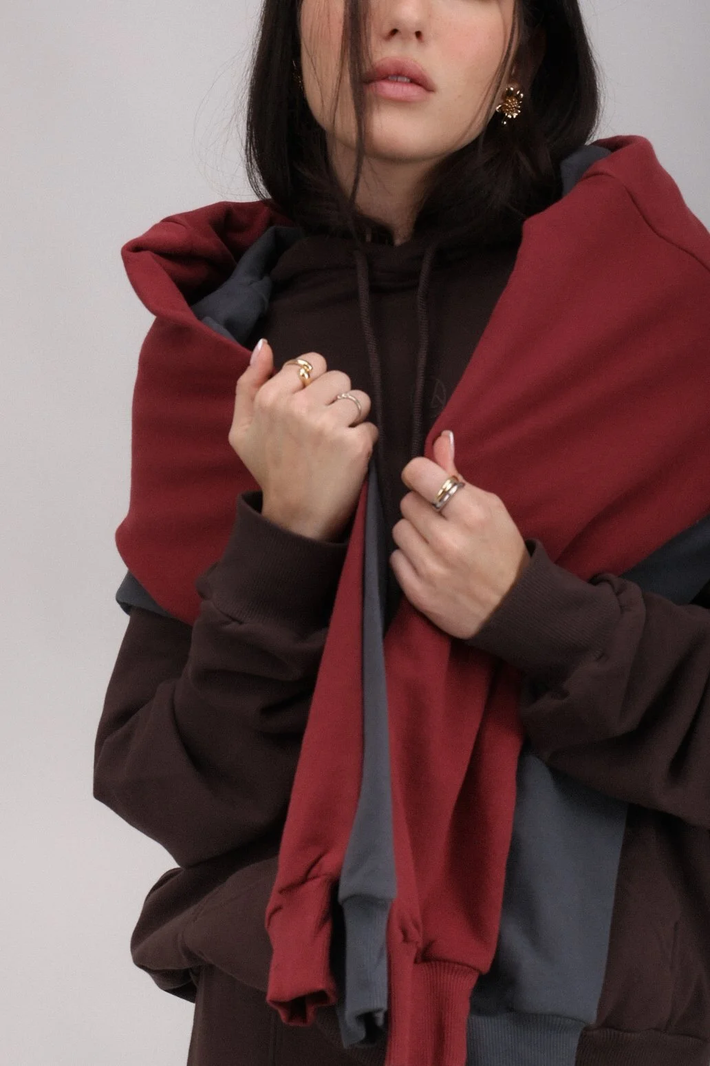A woman with dark hair wearing a multicolored jacket and jewelry, holding the front of her jacket with both hands.