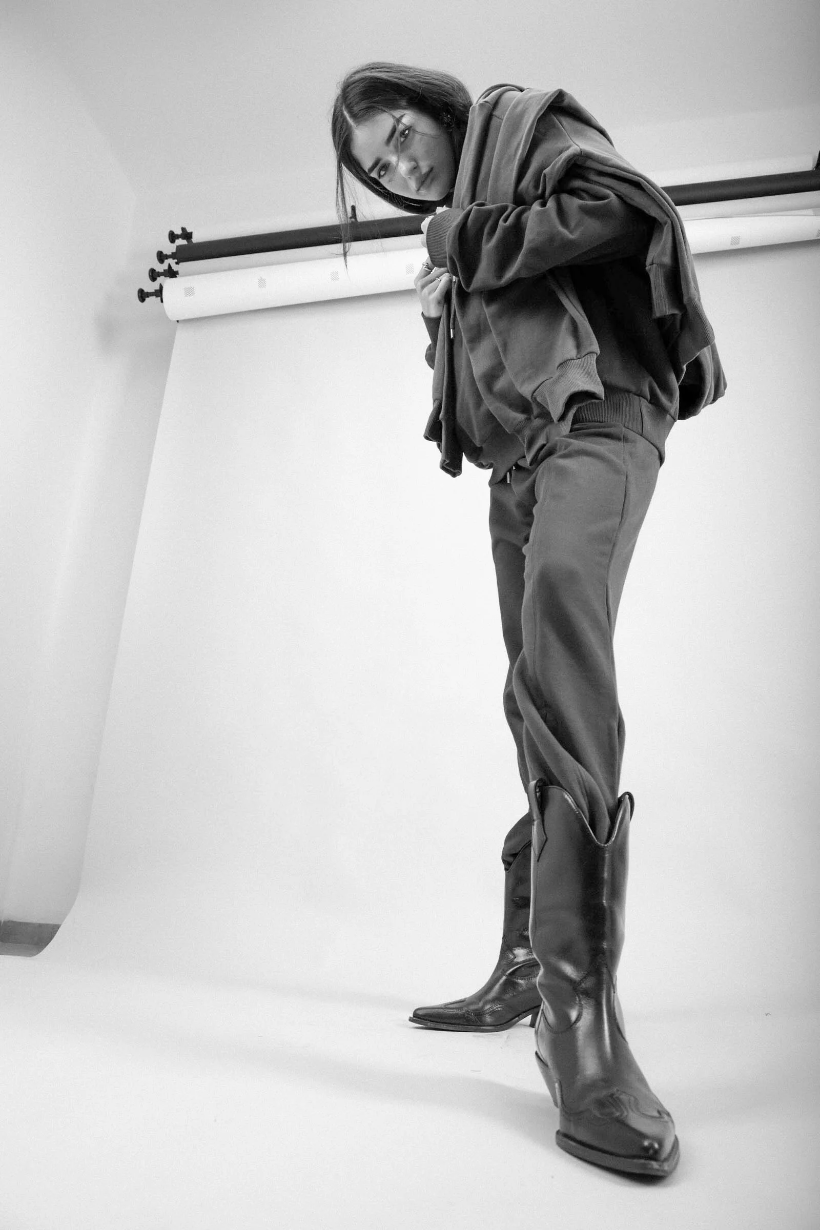 Black and white photo of a woman leaning against a wall with a photography backdrop frame. She is wearing glasses, a hoodie, loose pants, and shiny cowboy boots.