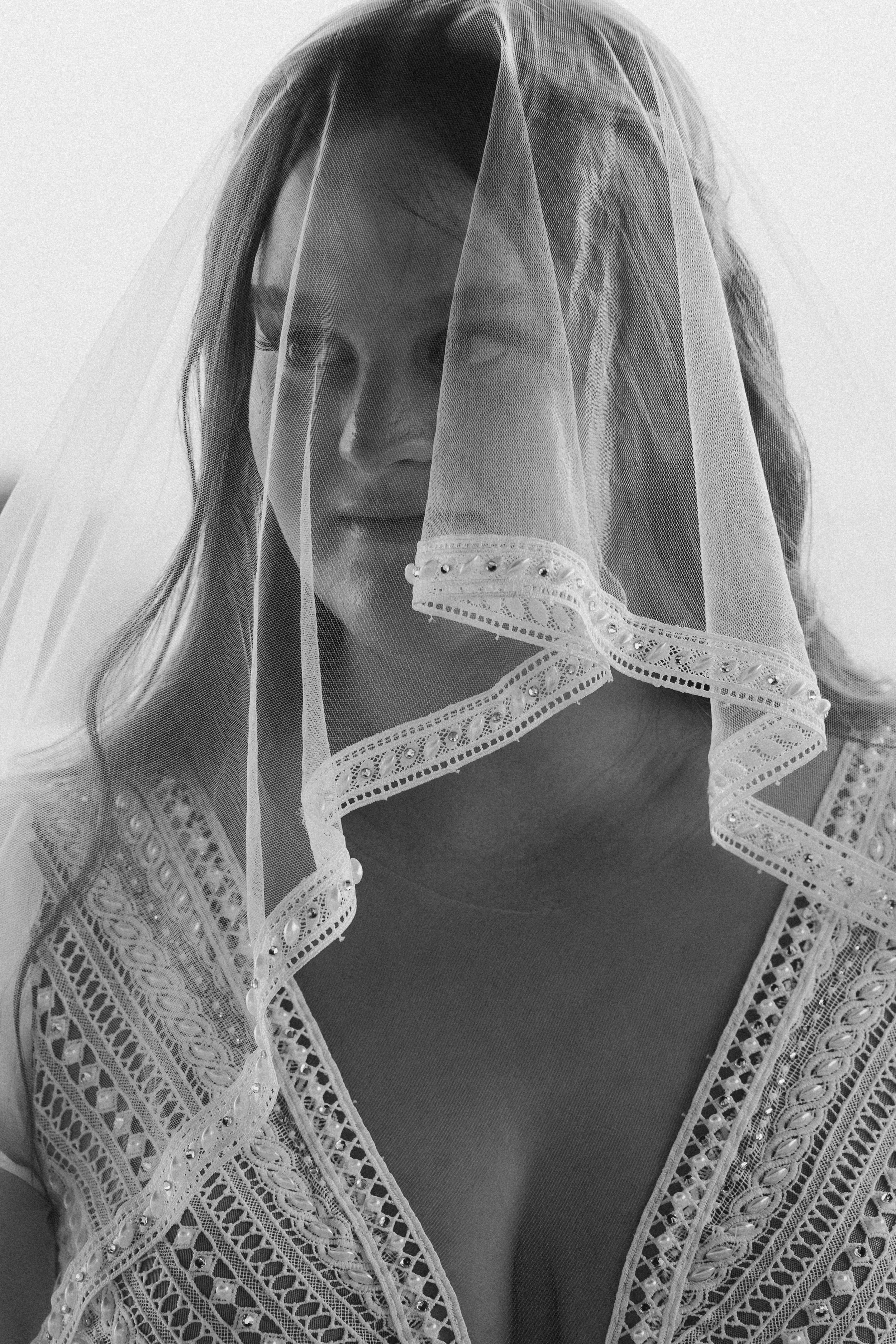 A woman with light hair partially covered by a veil or lace head covering, looking downward. She is wearing a lace or embroidered garment with intricate patterns.