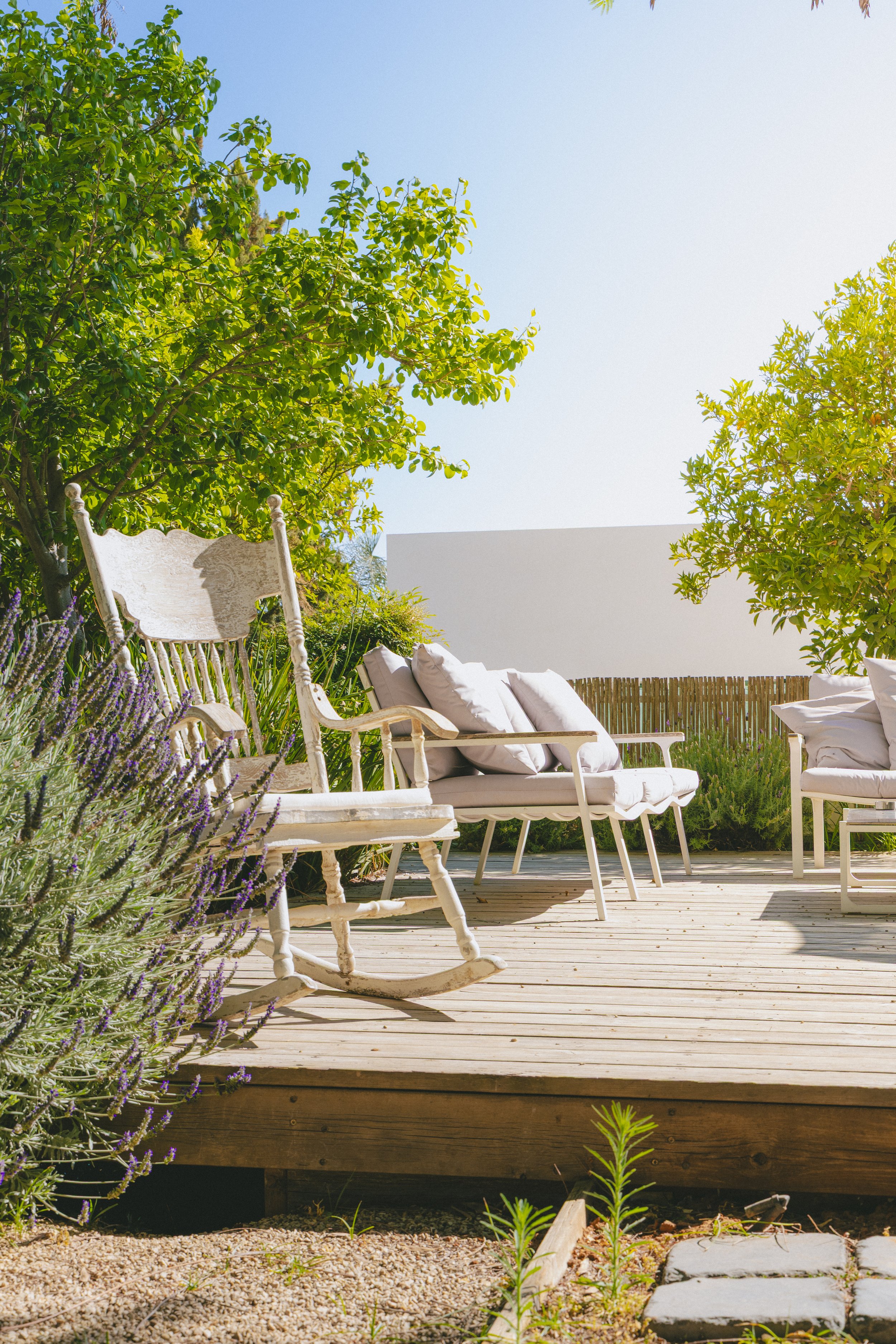 A wooden deck with white outdoor furniture, including a lounge chair with cushions, surrounded by greenery and purple flowers under a clear blue sky. athmosphere photoshoot, צילומי אווירה, צילומים לעסקים, תדמית לעסקים, צילומי חוץ