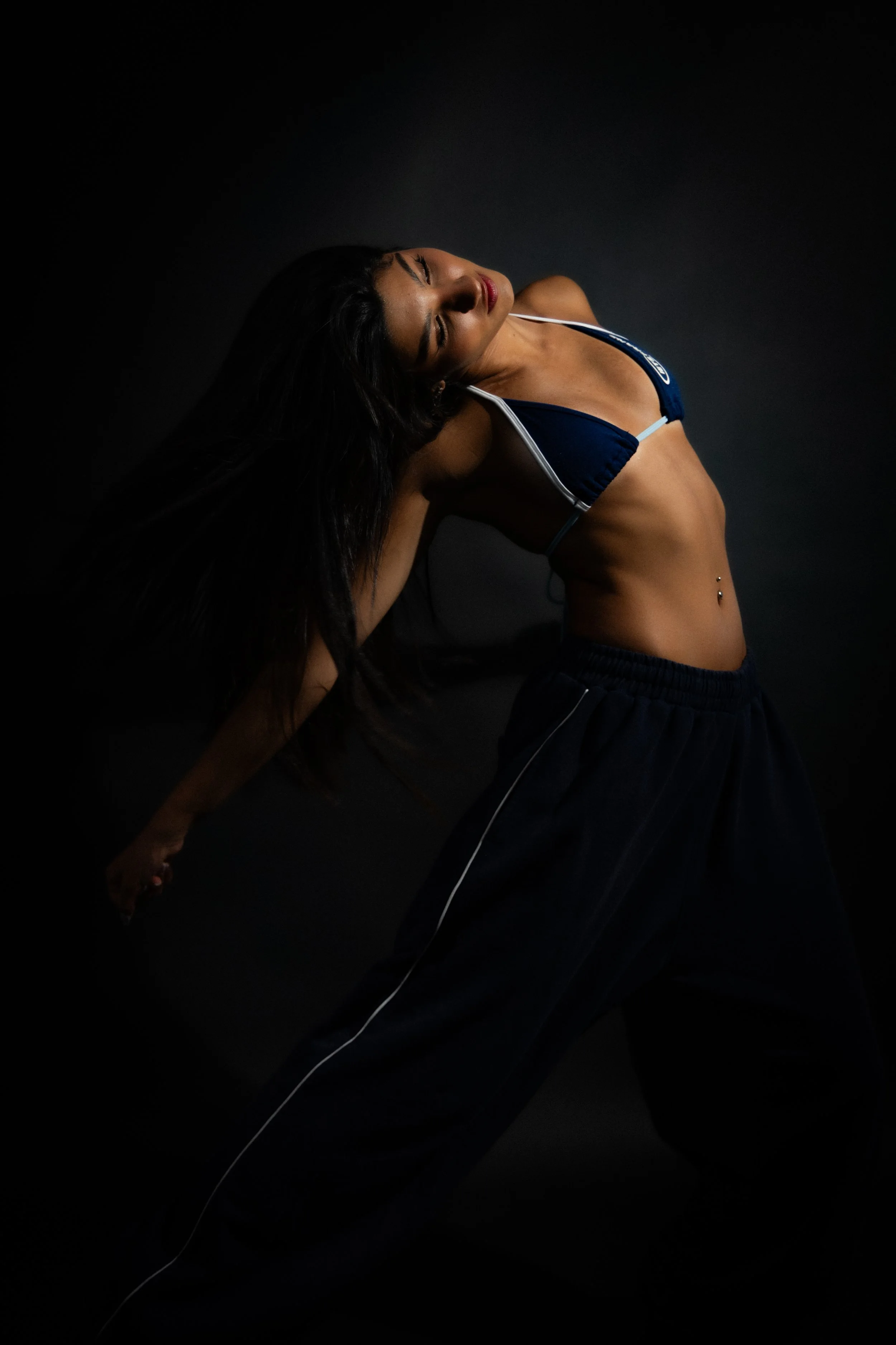 A woman with long black hair, wearing a navy blue sports bra and black pants, leans back with her eyes closed against a dark background.