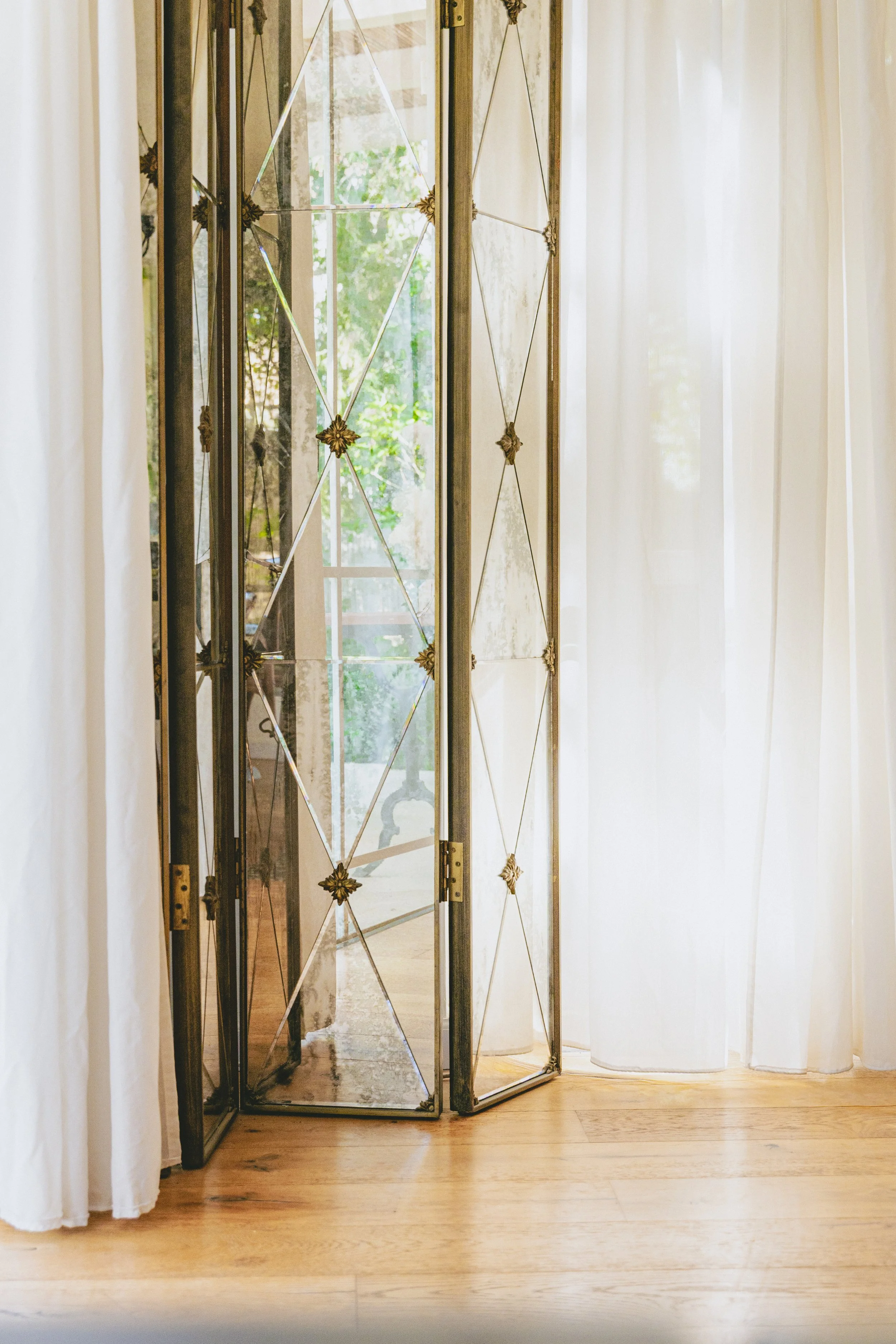 A folding decorative mirror with a metal frame and star-shaped accents stands in front of cream-colored curtains next to wooden flooring.