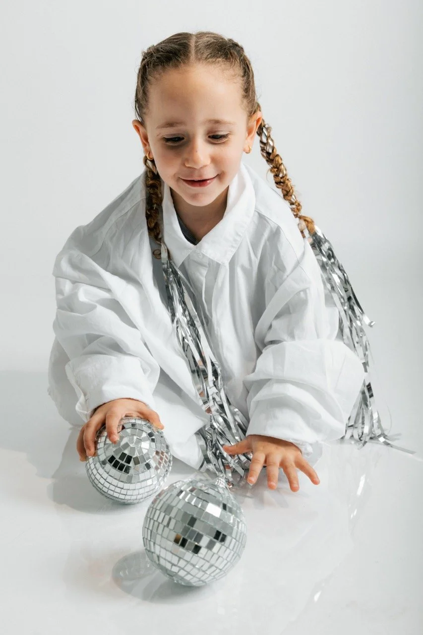 Young girl with braided hair wearing a white shirt with silver tinsel, playing with two mini disco balls on a white surface against a plain white background.