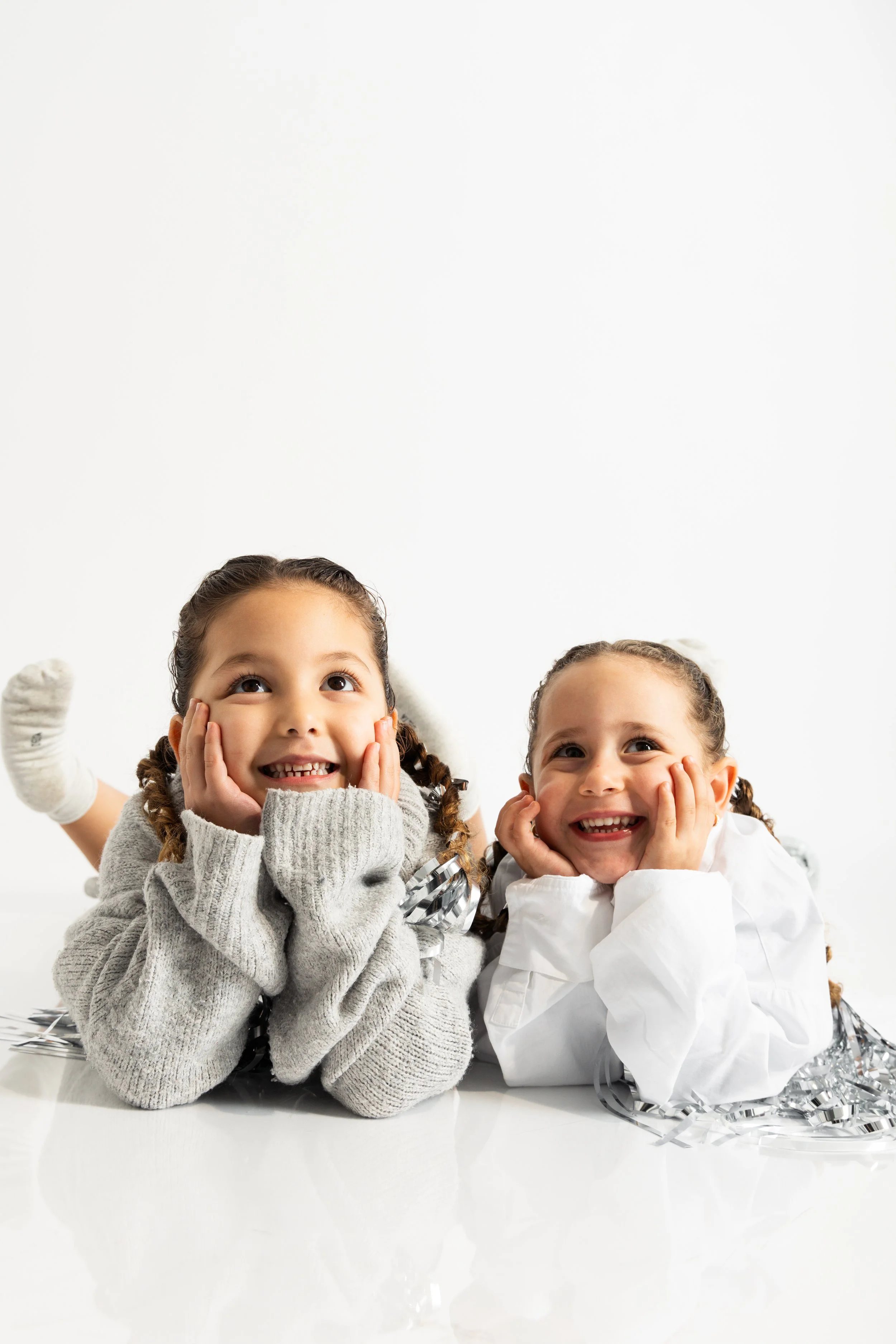 Two smiling young girls with braided hair, wearing cozy sweaters and makeup, lying on a white surface with silver tinsel decorations, against a plain white background.