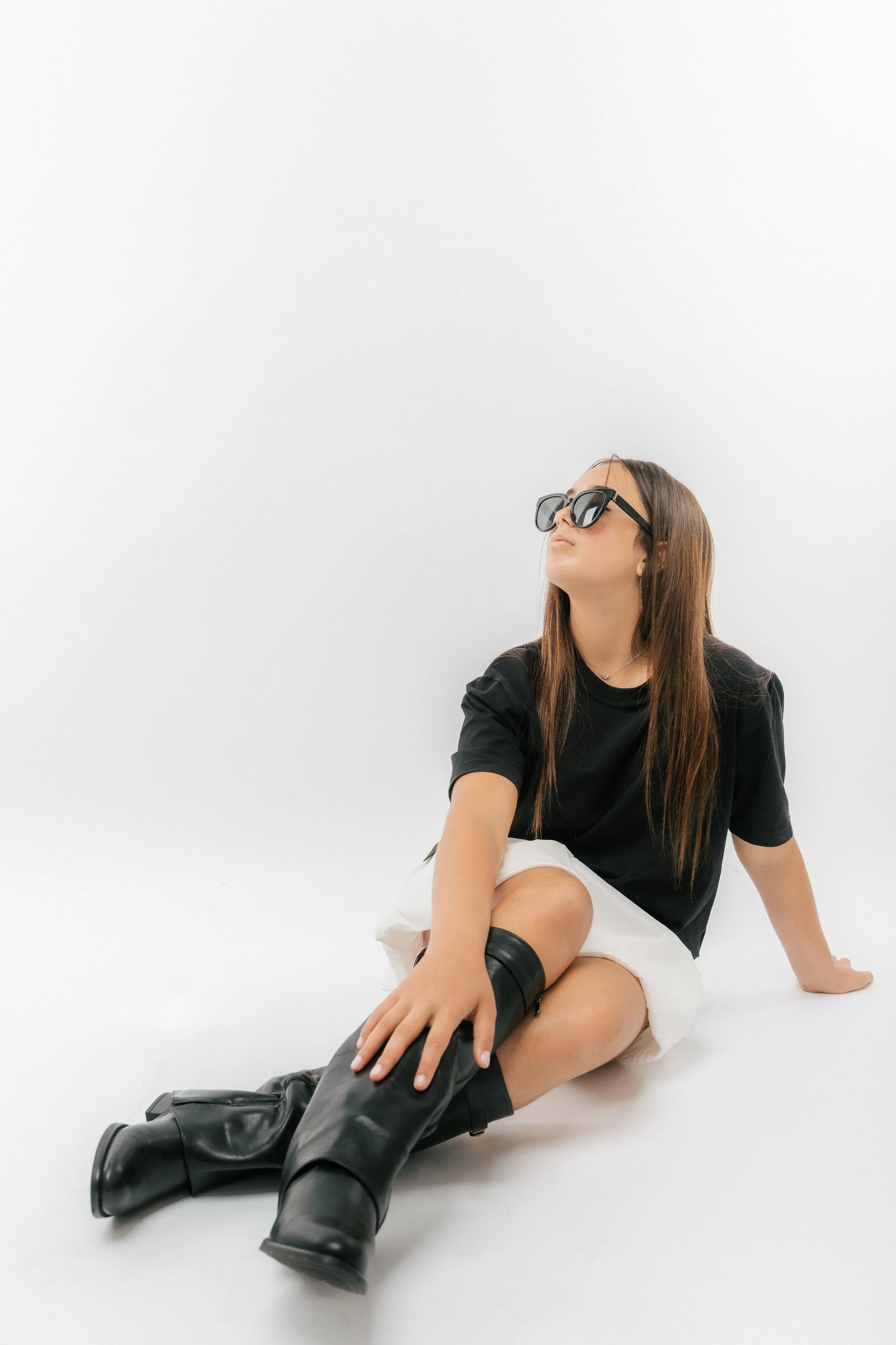 A young woman with long brown hair wearing black sunglasses, a black t-shirt, white shorts, and black knee-high boots, sitting on the floor against a plain white background, looking upward.