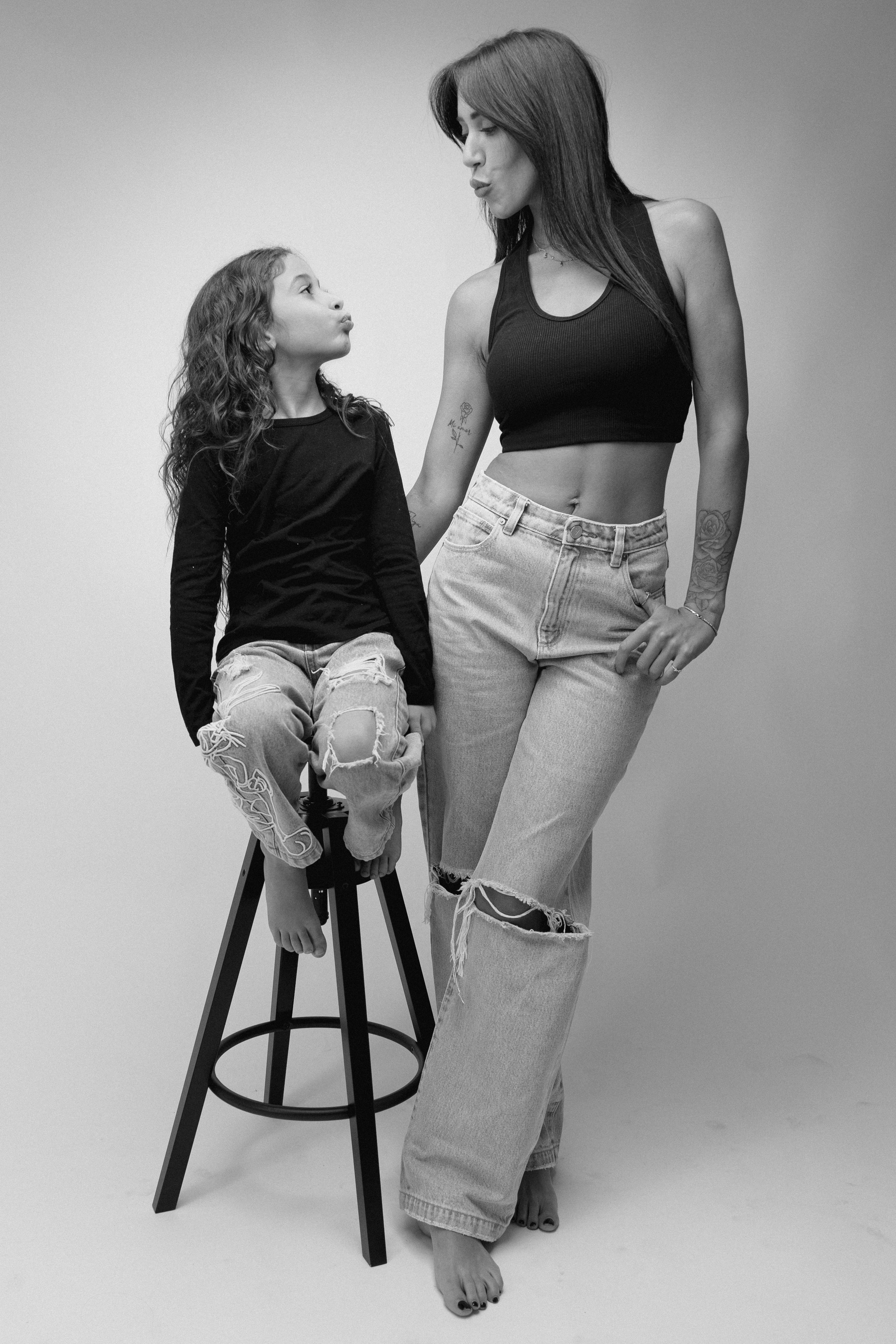 A woman and a young girl are standing and sitting respectively in a photo taken in a studio with a plain background. The woman is wearing a black crop top and ripped jeans, and the girl is wearing a black long-sleeve shirt and distressed jeans. The w