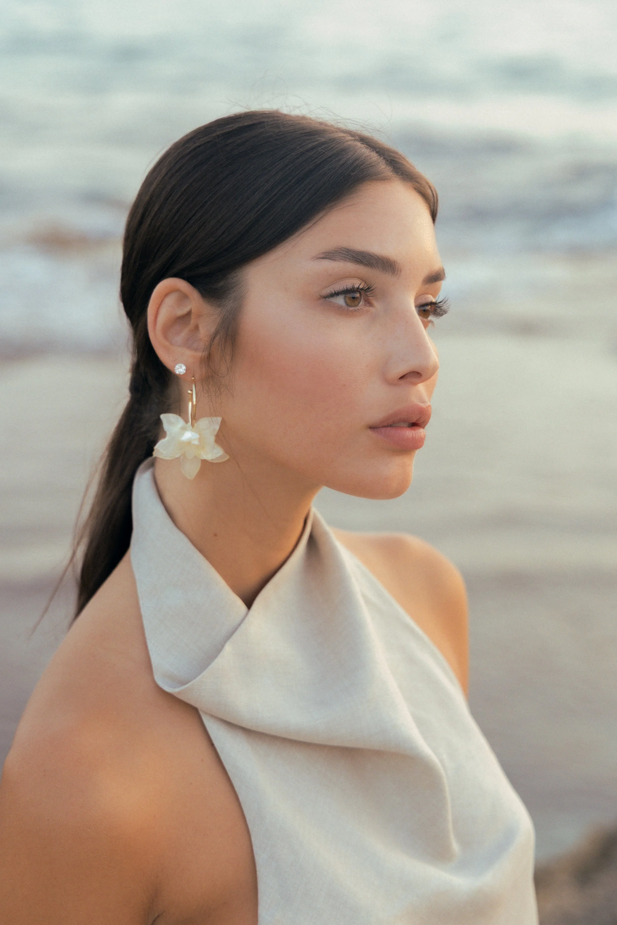 Close-up of a woman with dark brown hair styled in a sleek ponytail, wearing pearl earrings with flower pendants, looking to the right, near the beach with waves in the background. fashion photography. צילומי אופנה, צילום למותגים, דוגמנית. פורטרייט