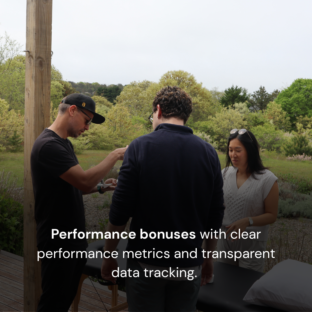 Three people standing outdoors, two men and one woman, engaged in conversation or looking at something, with trees and greenery in the background.