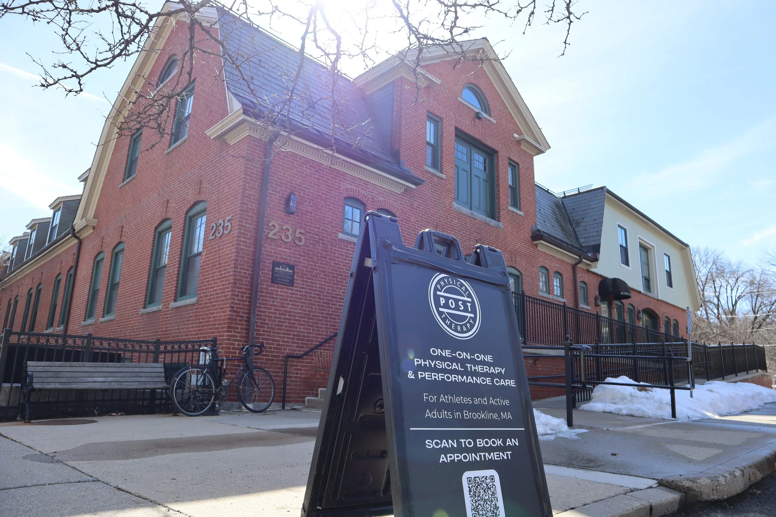 A brick building with gray roof and large windows, a sign in front advertises physical therapy services, bicycles parked nearby, and patches of snow on the sidewalk.