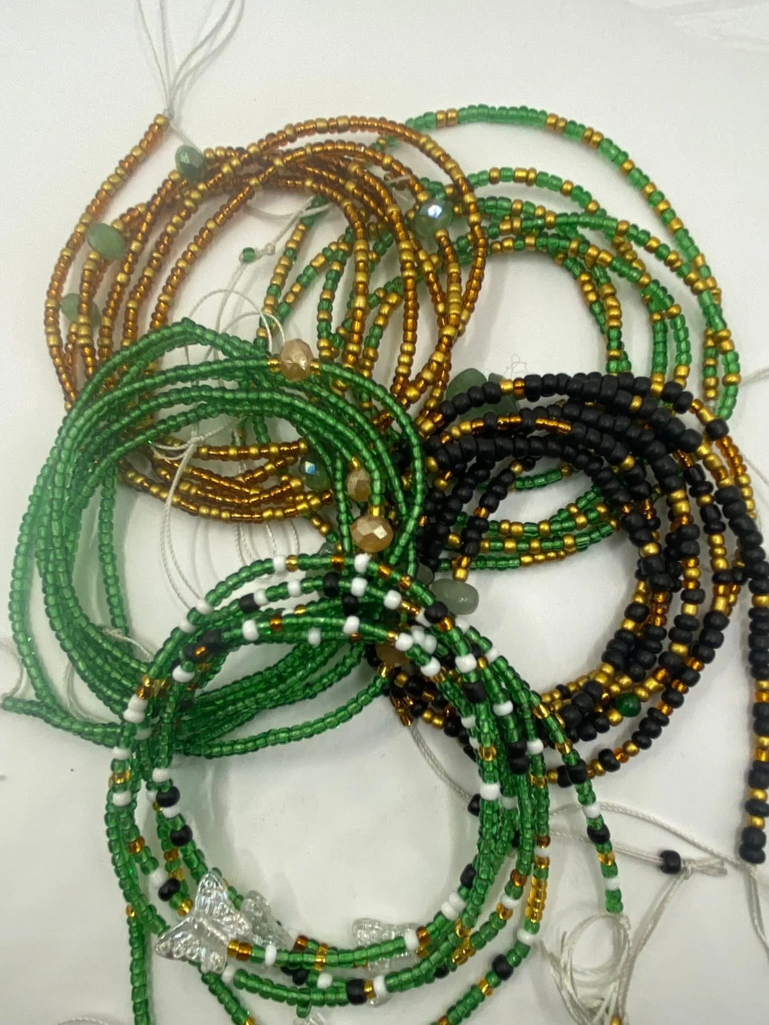 Colorful beaded necklaces in black, green, gold, white, and clear beads, with some featuring star-shaped pendants, arranged on a white surface.
