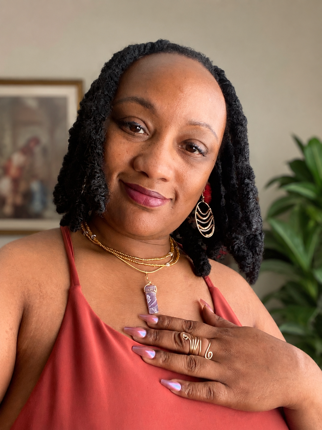A woman with short black curly hair and jewelry, wearing a red top, looking at the camera with her hand on her chest, in a room with a plant and a framed picture in the background.
