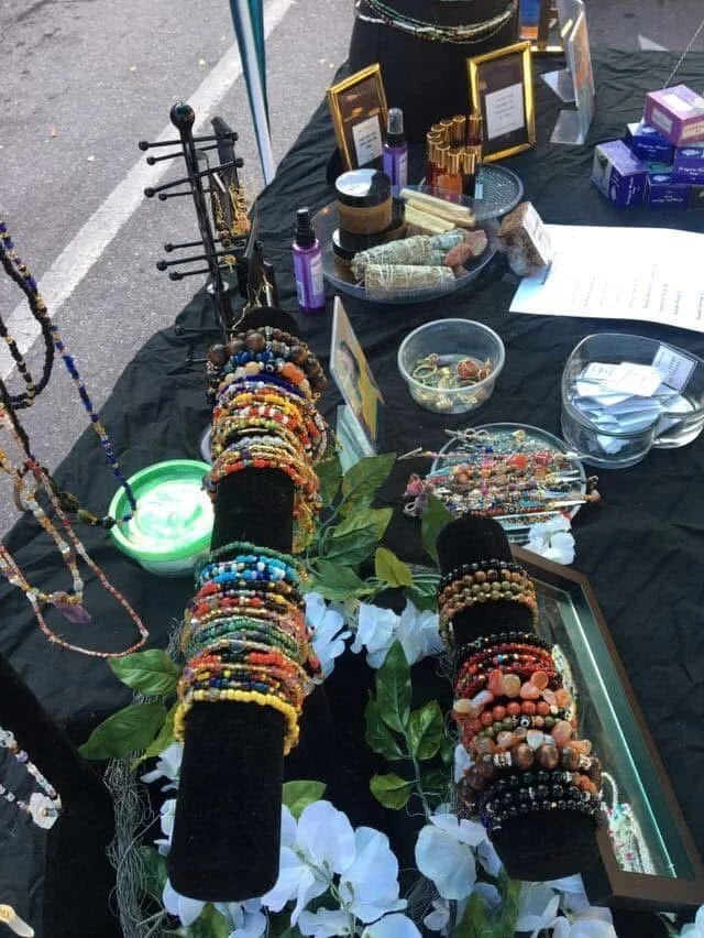 Jewelry display with numerous colorful bracelets and necklaces, set on black fabric with green leaves and white floral accents, at an outdoor market stall.