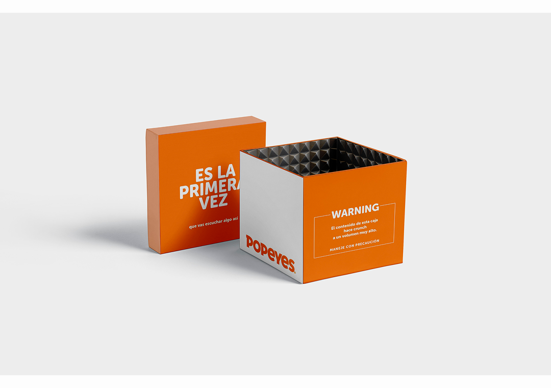Cubo de cartón de color naranja y blanco con la marca Popeyes, una tapa con un mensaje y un interior metálico antideslizante, en un fondo gris claro.